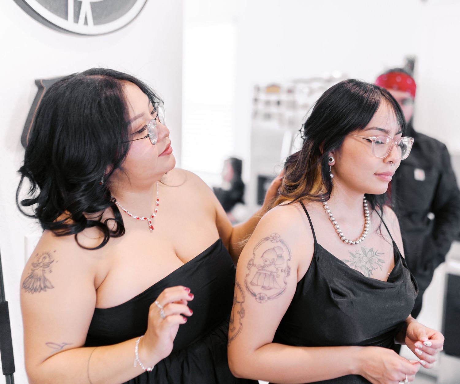Two people in black spaghetti strap dresses with tattoos and necklaces enjoying a casual moment together.