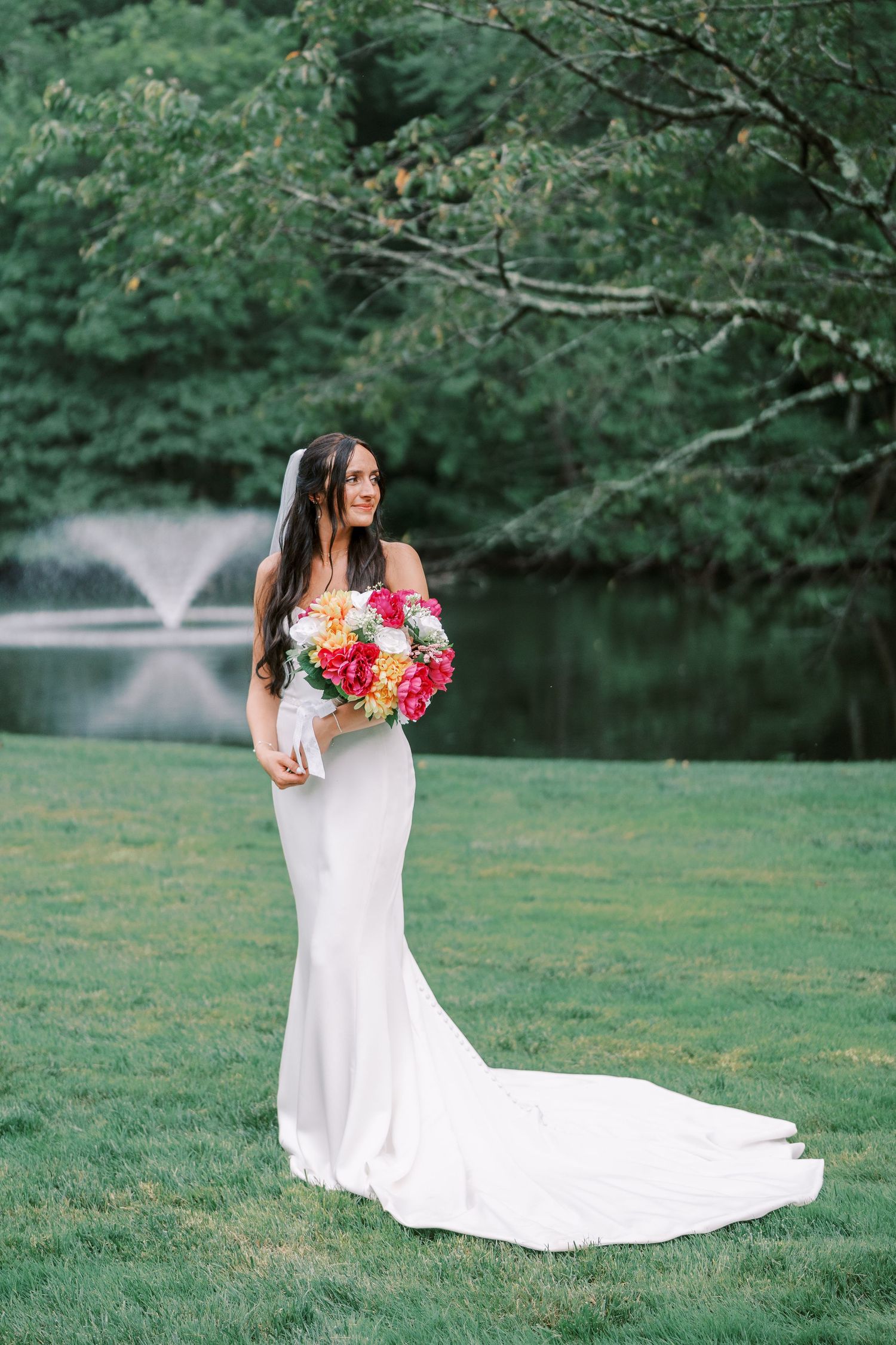 A bride in a flowing white gown holds a vibrant bouquet amid lush greenery and a fountain in the background.