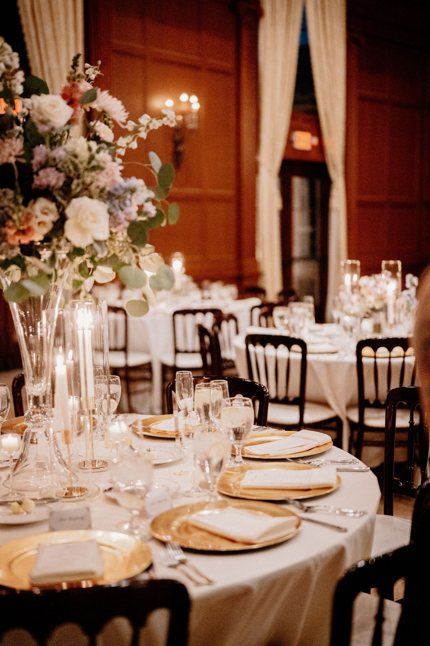 Elegant wedding reception tables with white linens, gold charger plates, and tall floral centerpieces in a warm-lit ballroom.