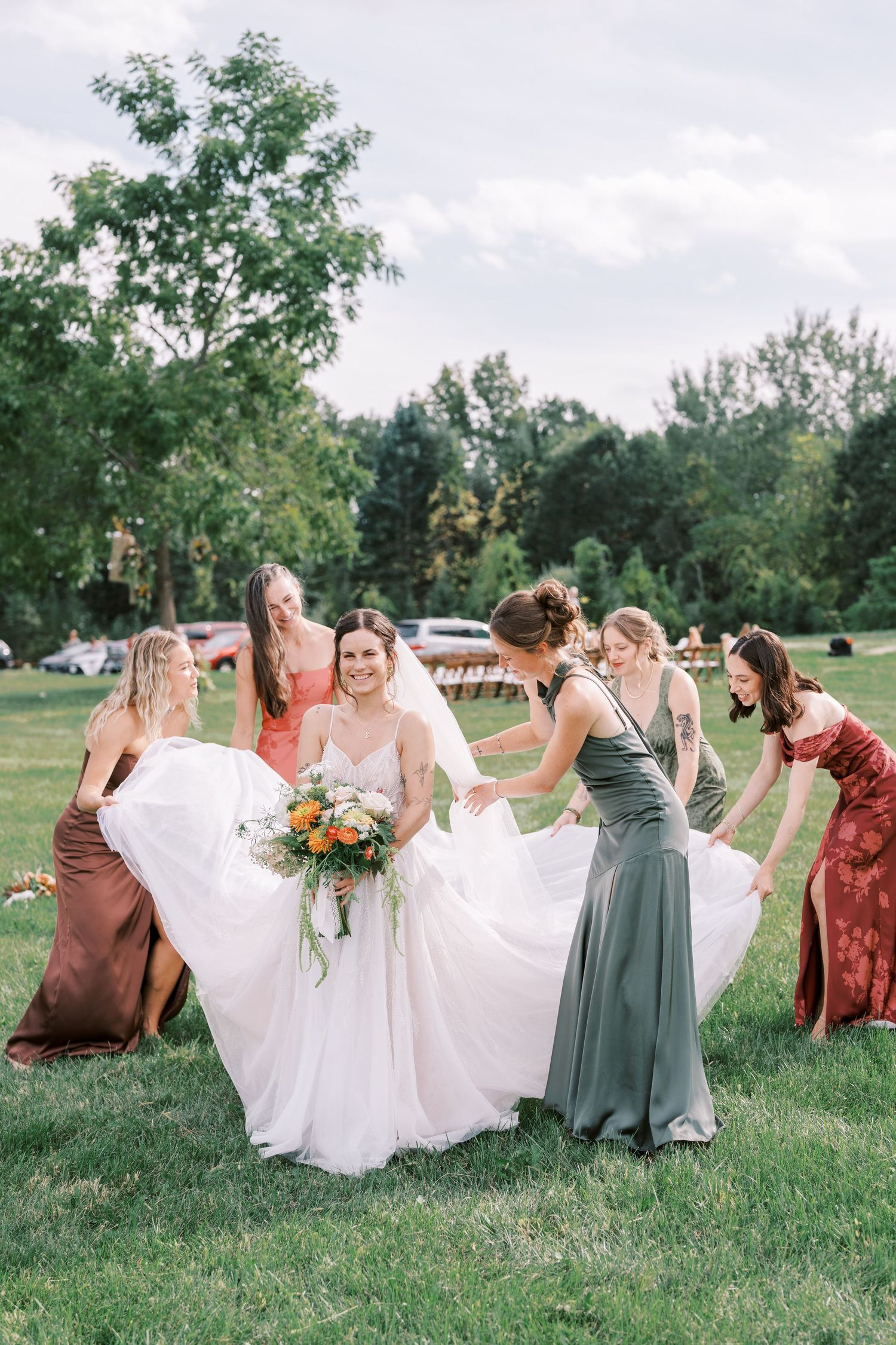 Bridesmaids in coral and sage dresses help arrange wedding dress on grass.