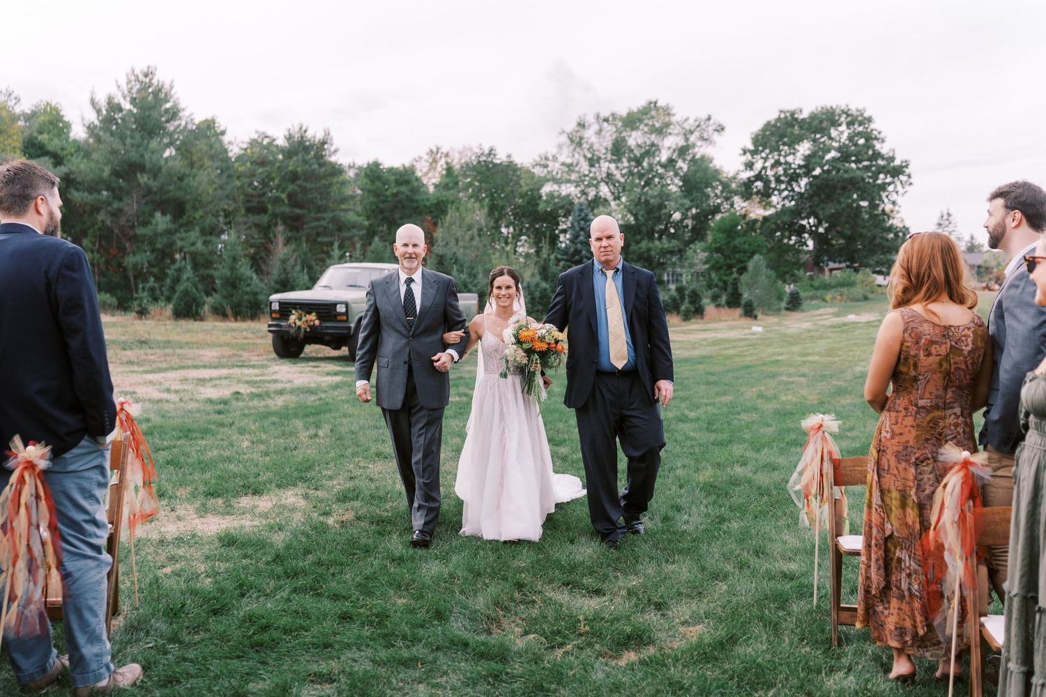 Wedding guests gather outdoors on a grassy hillside during a rustic countryside celebration.
