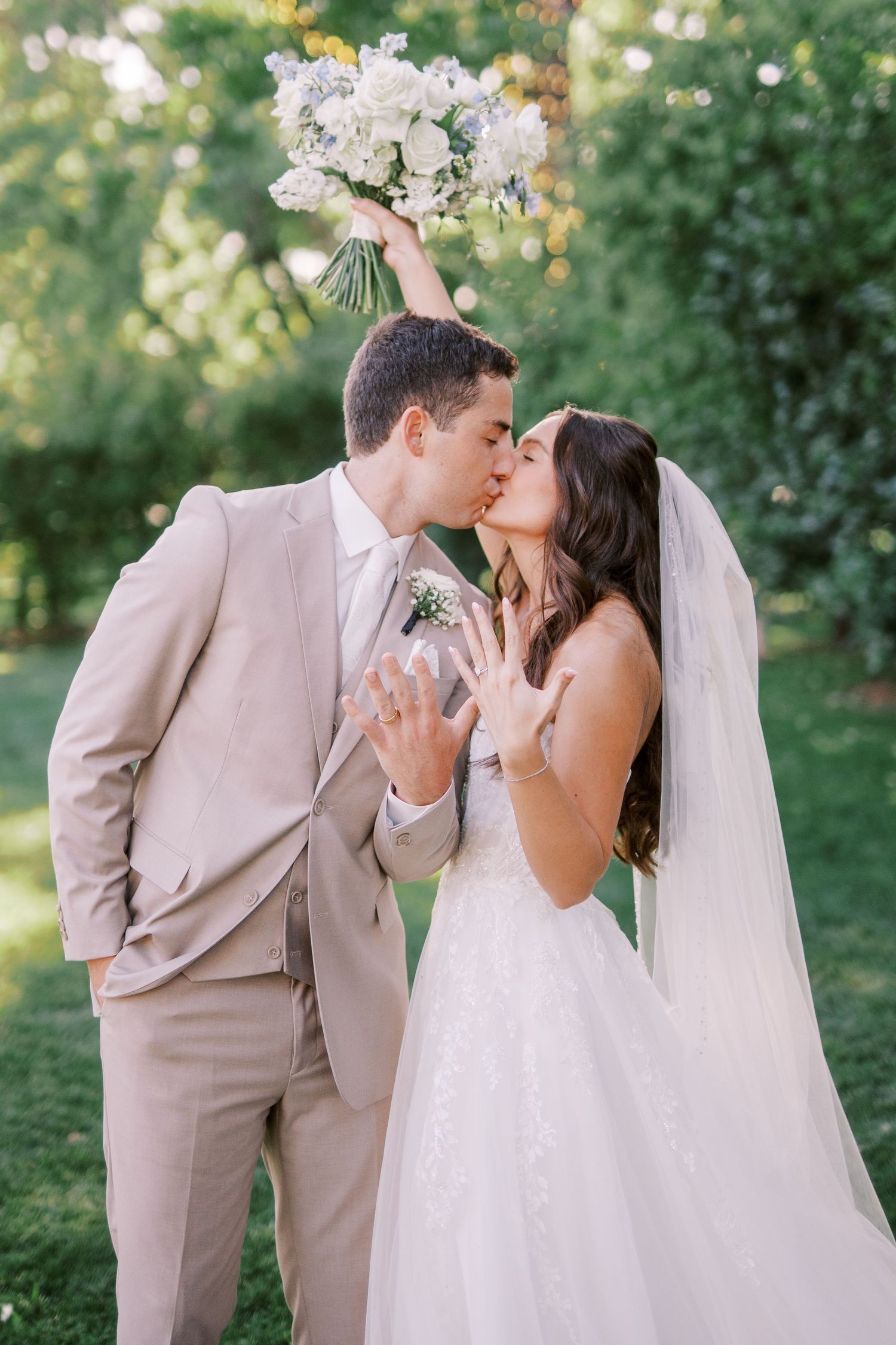 A couple in wedding attire outdoors with a tan suit and white dress share romantic kisses in a garden setting.