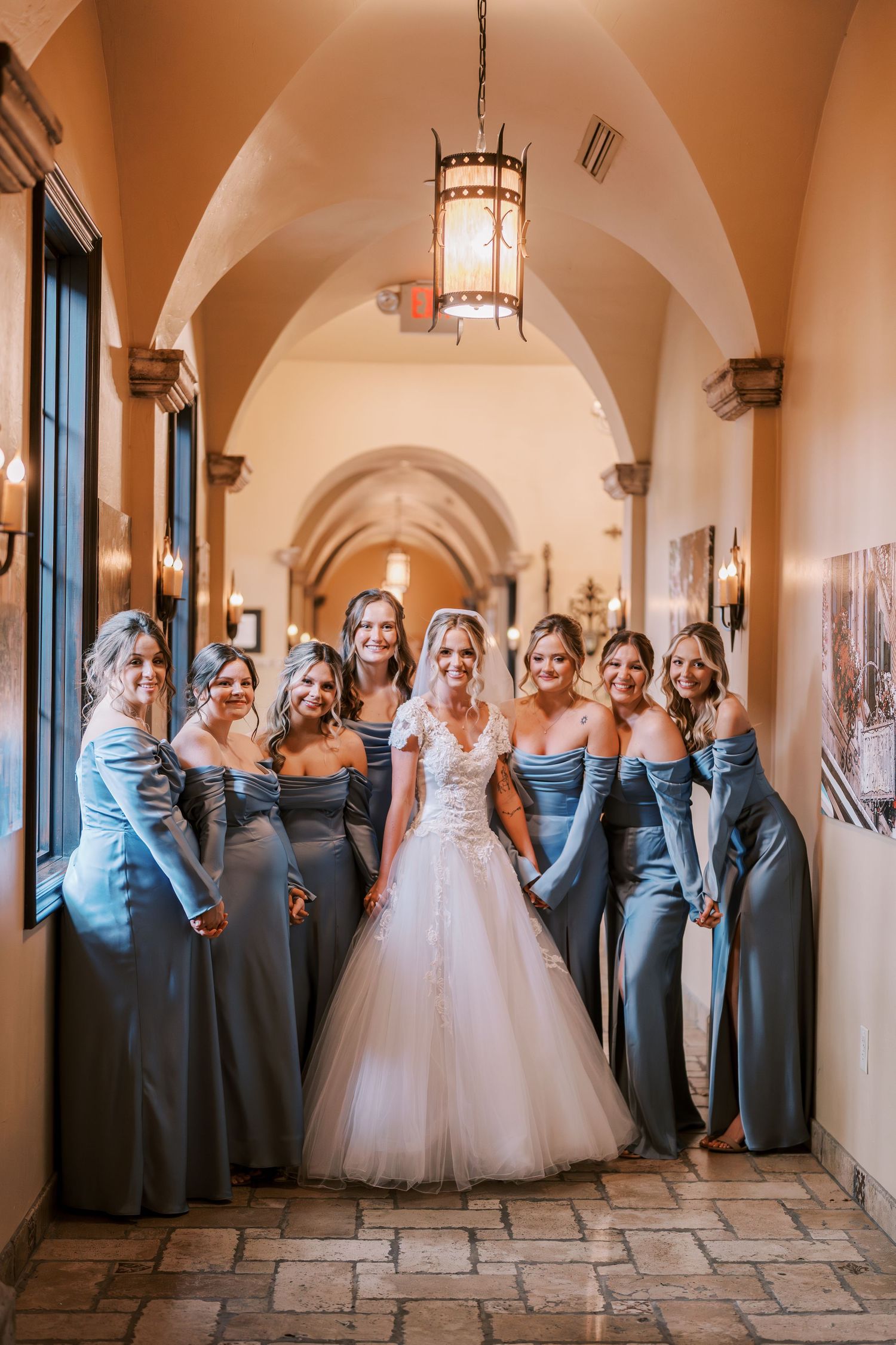 Wedding party wearing blue and gray dresses poses together in an elegant arched hallway with stone floors and pendant lights.