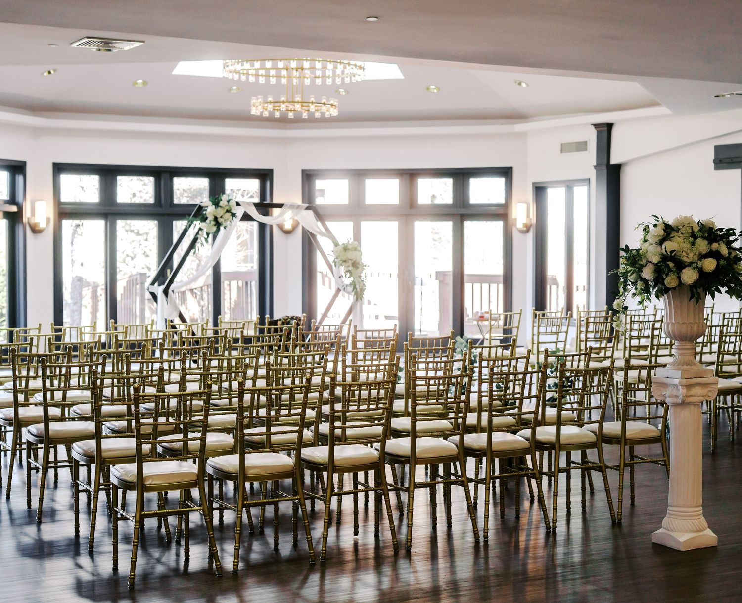 An elegant wedding ceremony space with rows of gold chairs and white floral arrangements under a crystal chandelier.