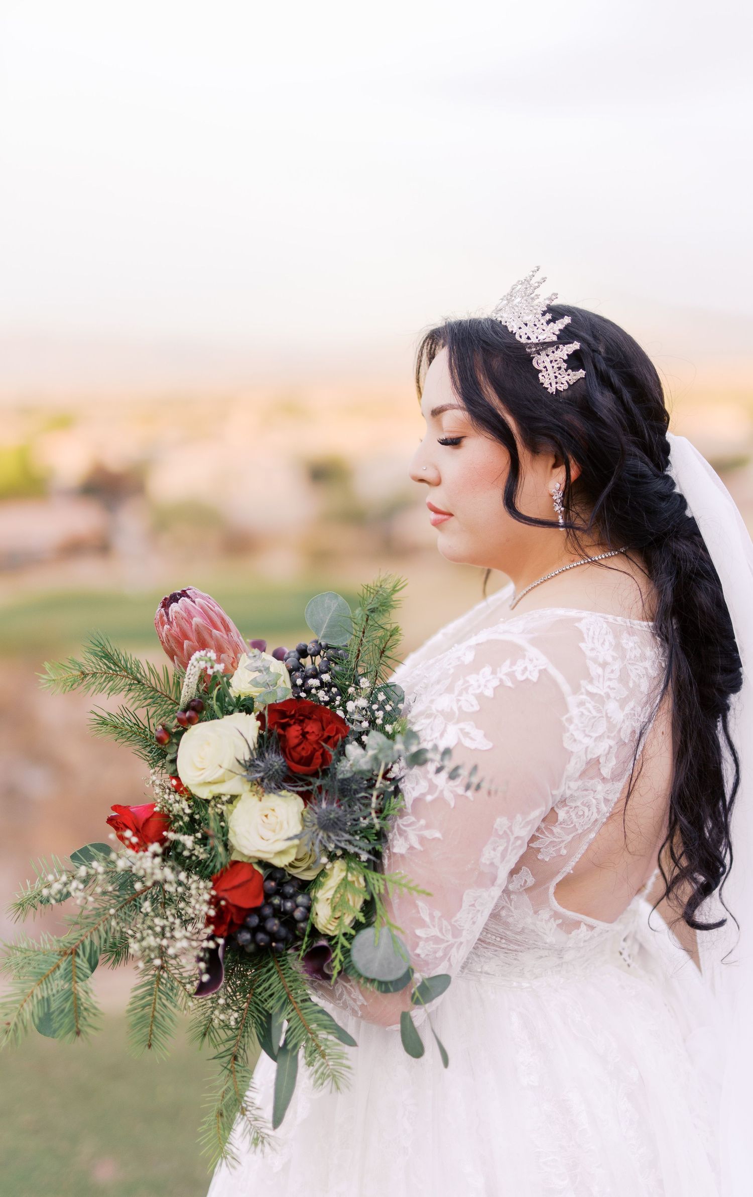Elegant white lace wedding dress and crystal crown paired with romantic winter bouquet featuring red and white roses at sunset.