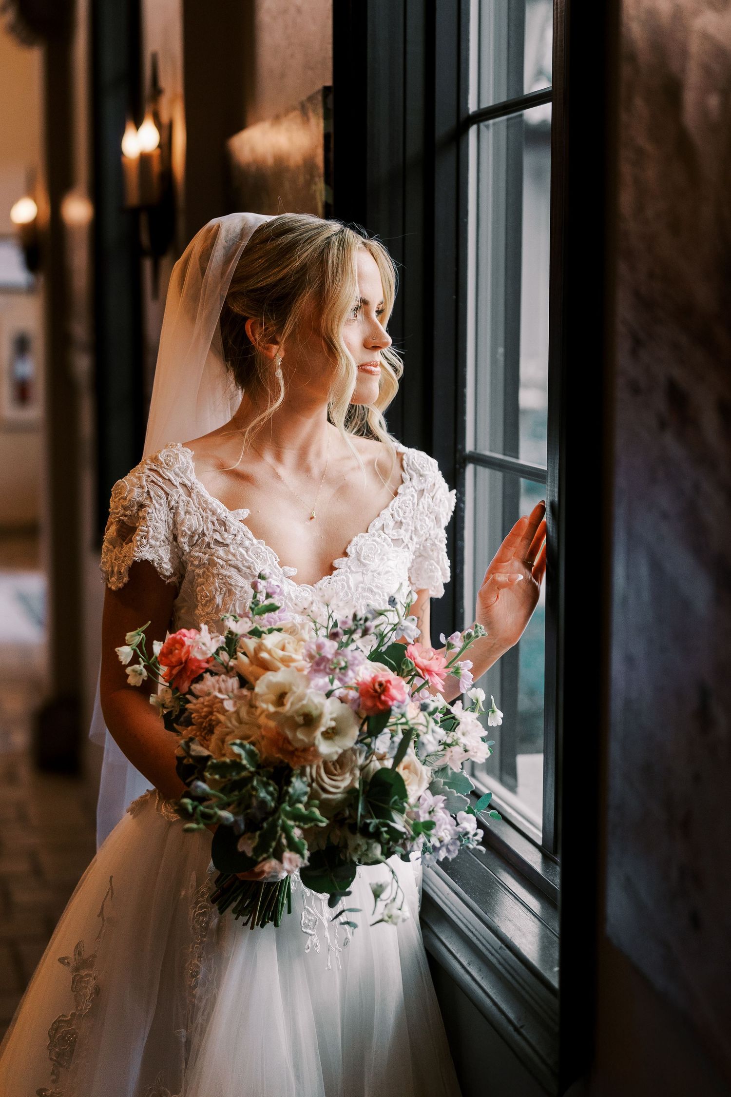 Bride in lace dress with floral bouquet gazes out window in romantic wedding portrait series.