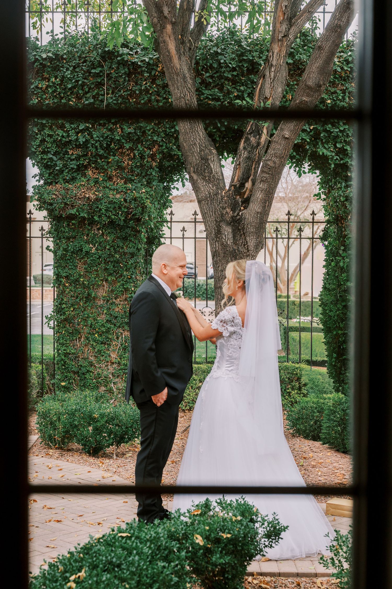 Wedding couple shares a private first look moment in a garden courtyard framed by window panes and lush greenery.