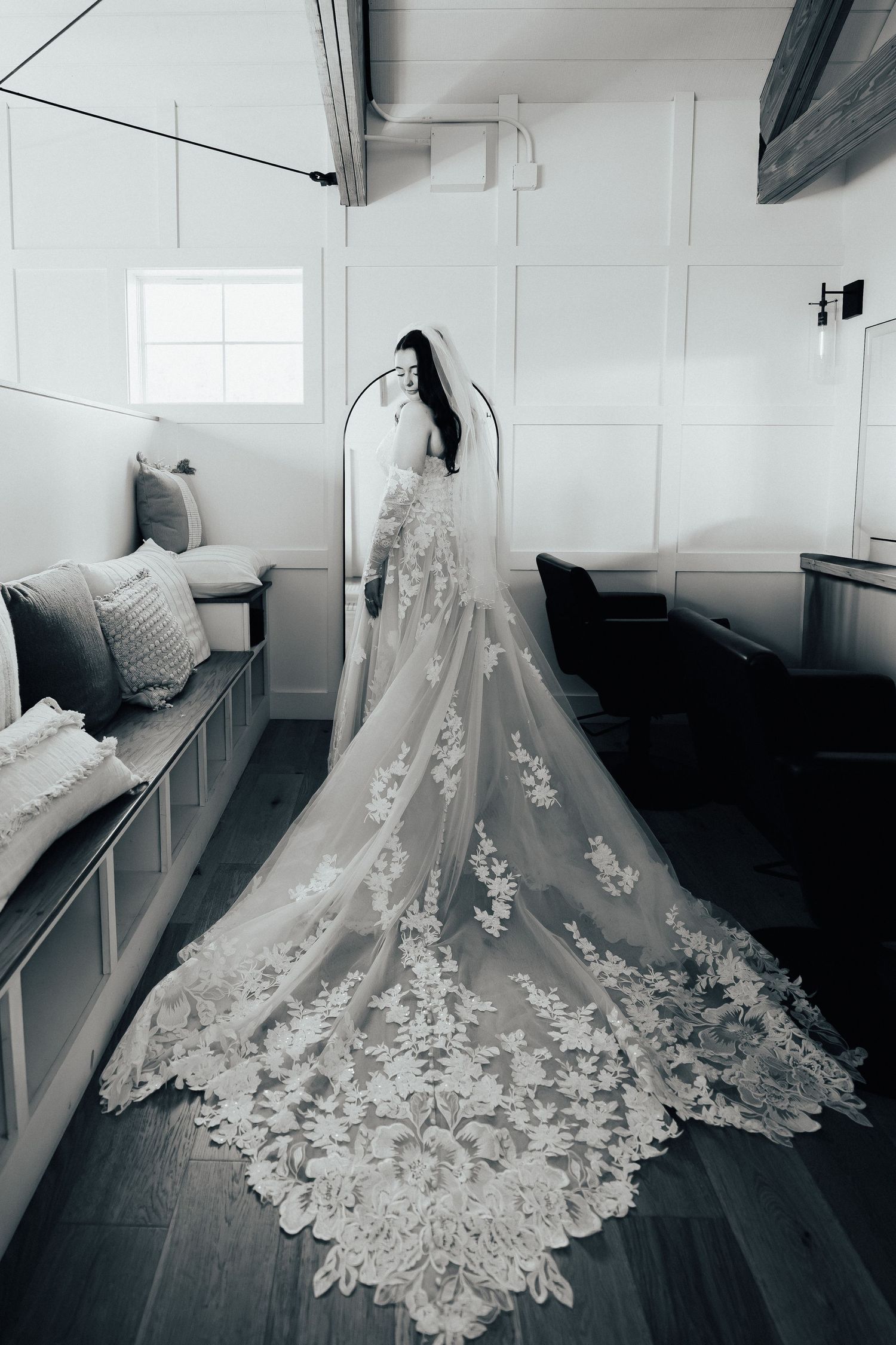 A stunning white wedding dress with intricate lace details and long cathedral train displayed on a hanger in a simple room.