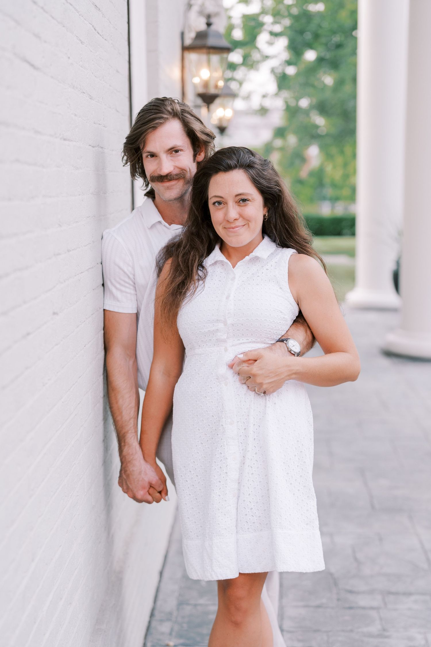 A couple in white outfits shares a playful moment against a light colored wall.