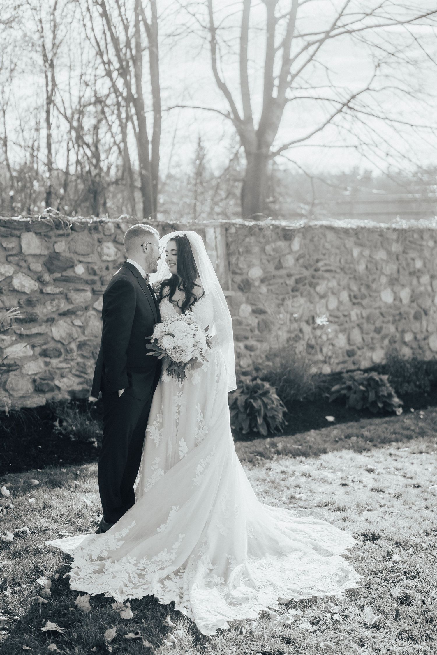 A black and white wedding photo of a couple standing by a rustic stone wall with bare winter trees in the background.