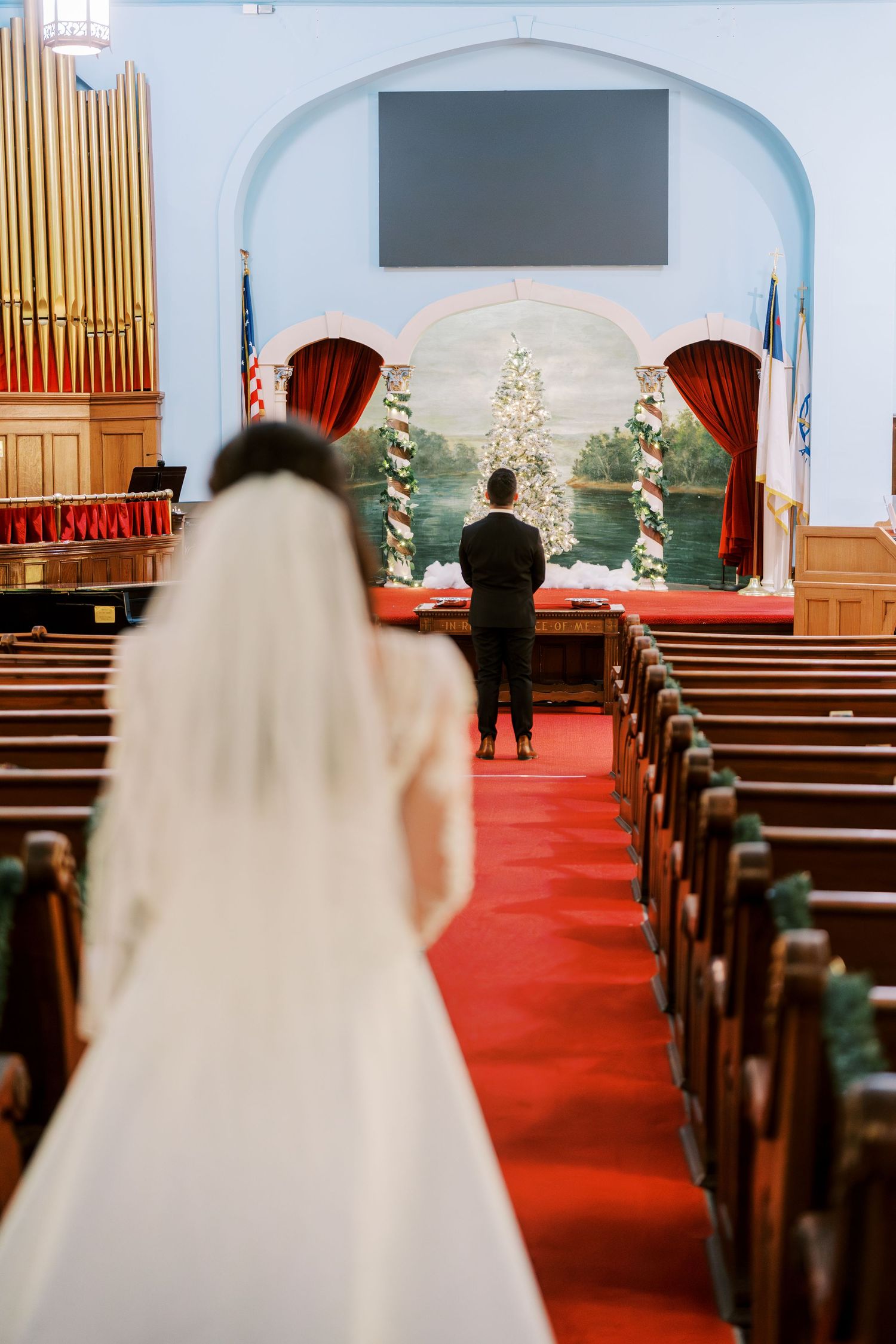 A bride in white walks down a church aisle decorated with Christmas trees and red carpet.