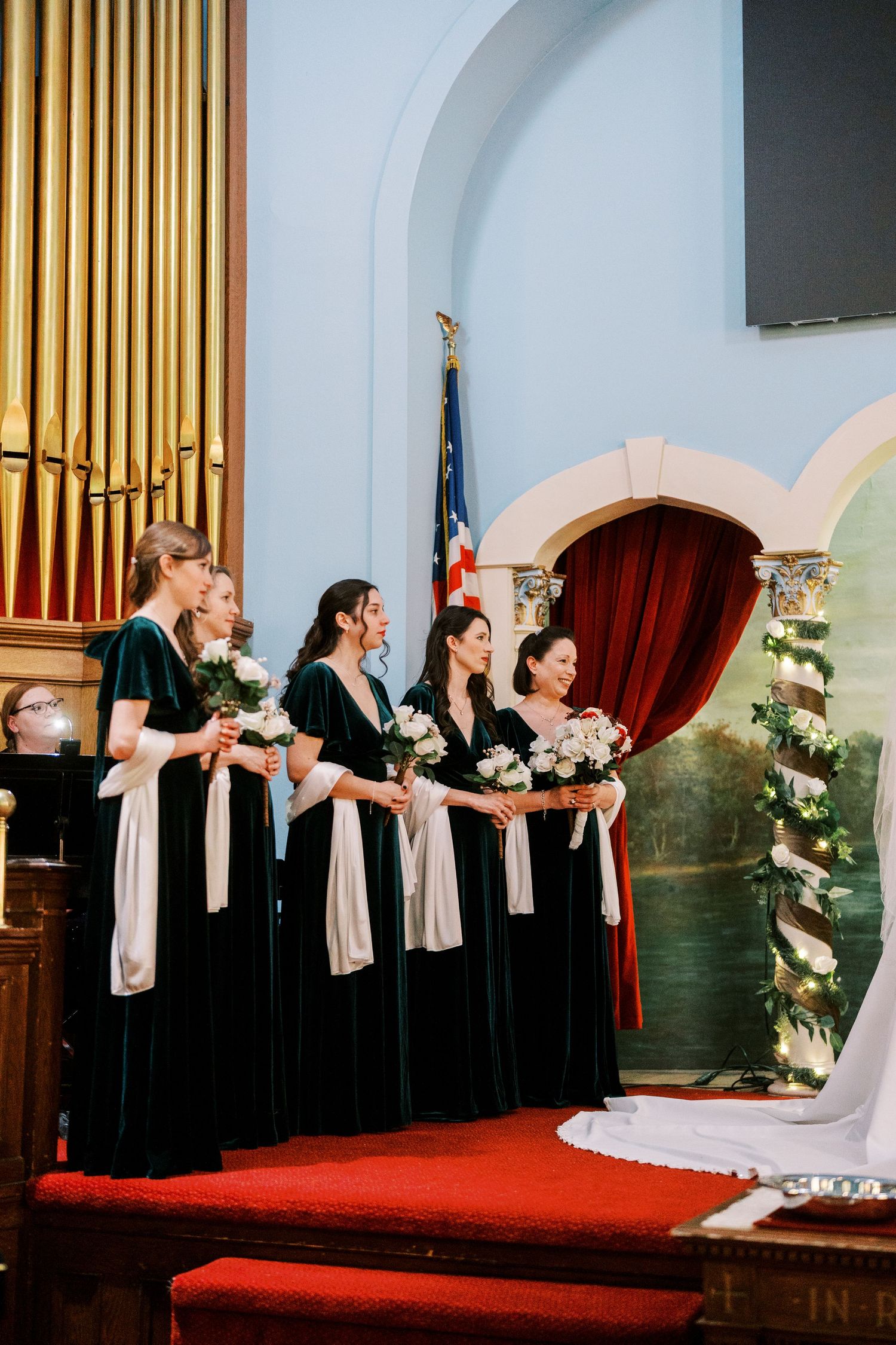Bridesmaids in black dresses hold bouquets while standing on church altar steps.