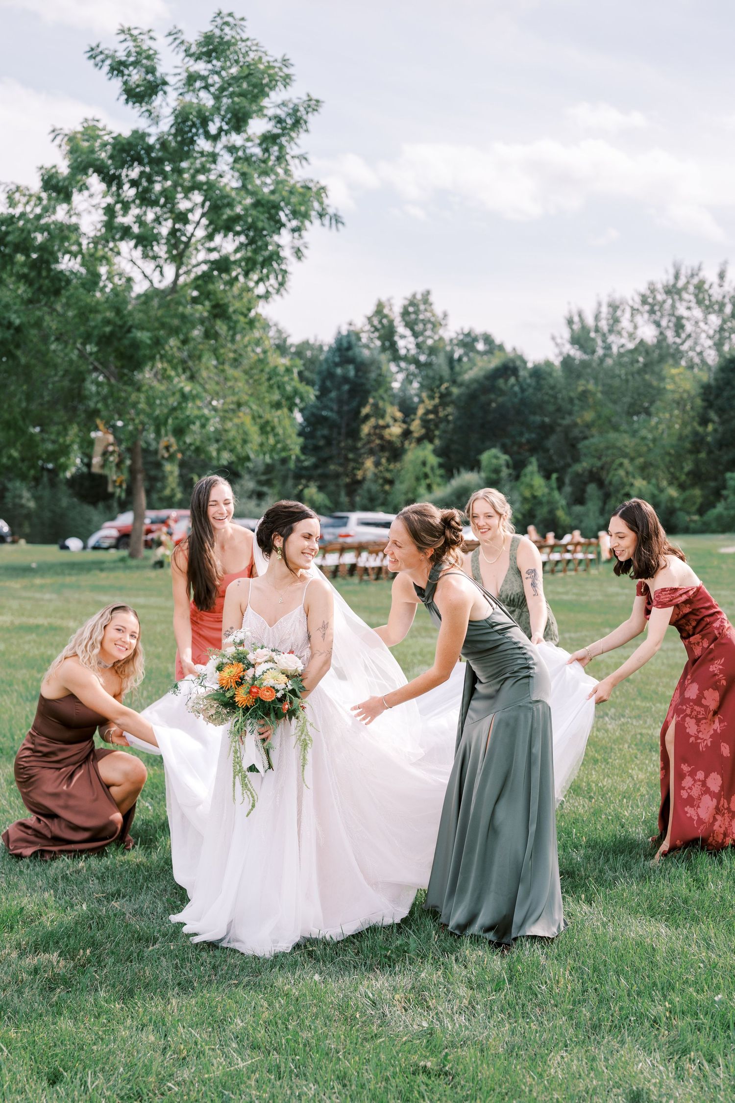 Wedding party in coordinating autumn-colored dresses sits playfully on grass during outdoor photo session.