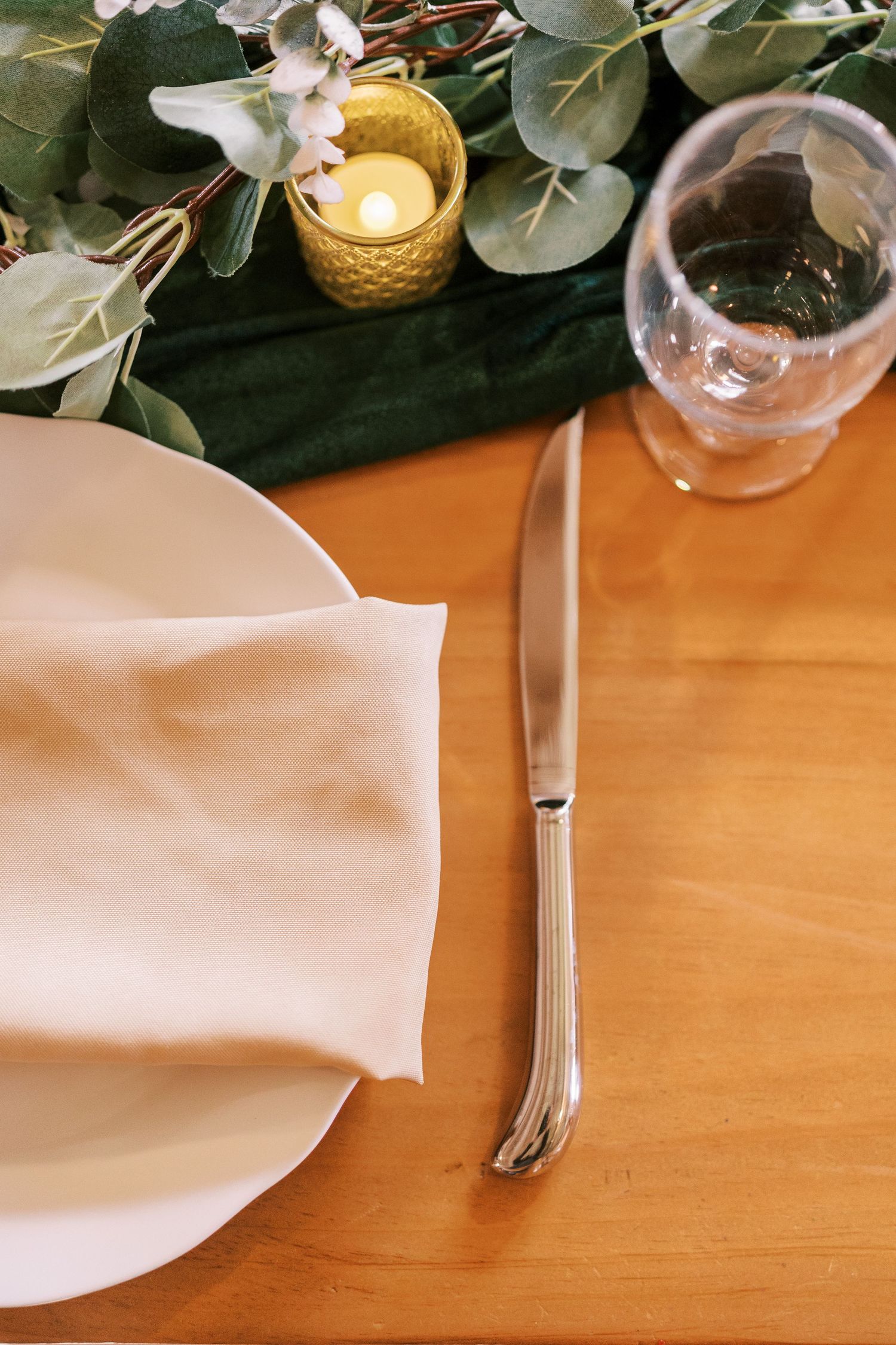 Sequence of white napkin folding steps shown on wooden table with decorative greenery and candles in background.