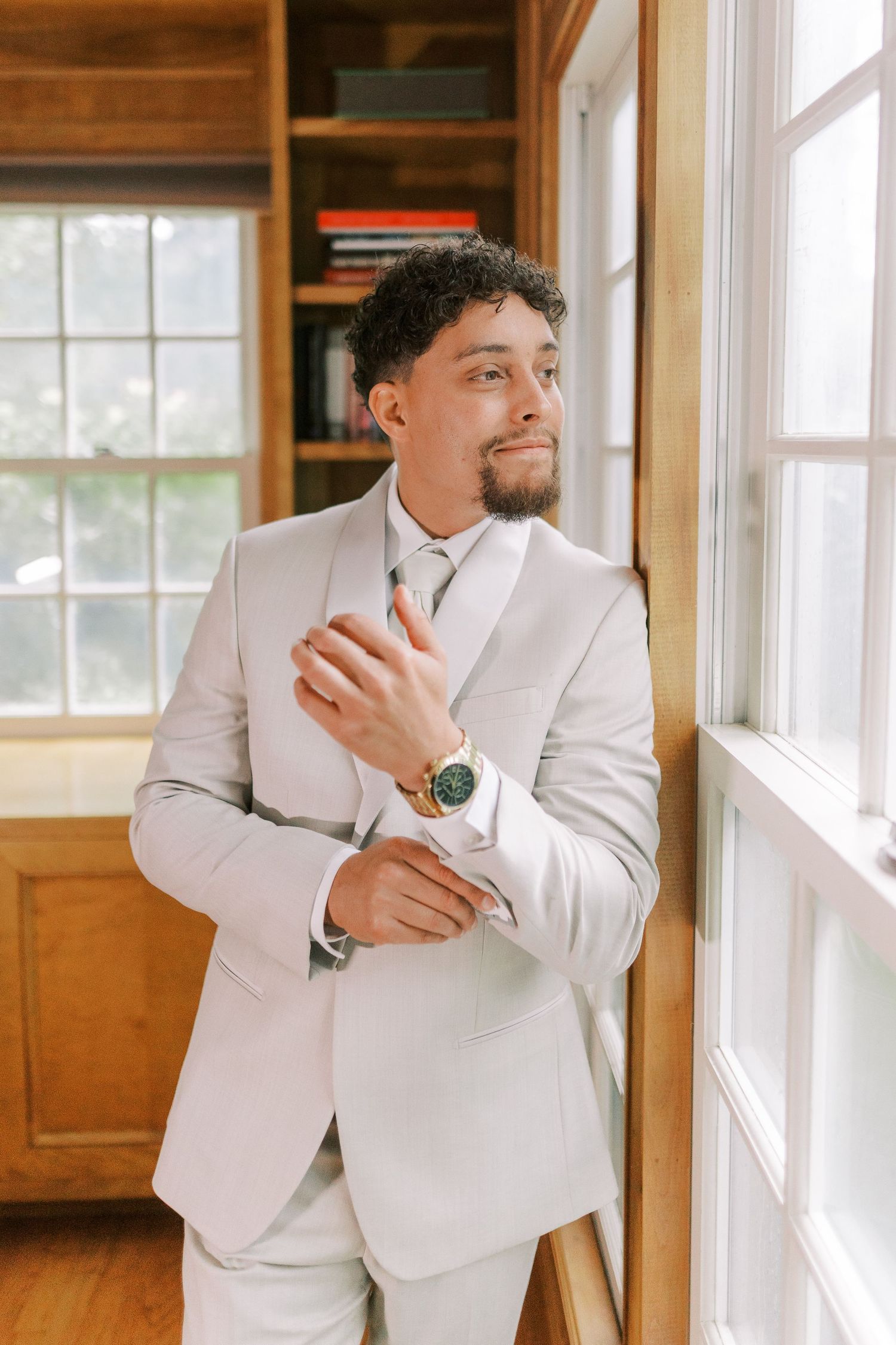 Groom in a light gray suit adjusting cufflinks by a window.