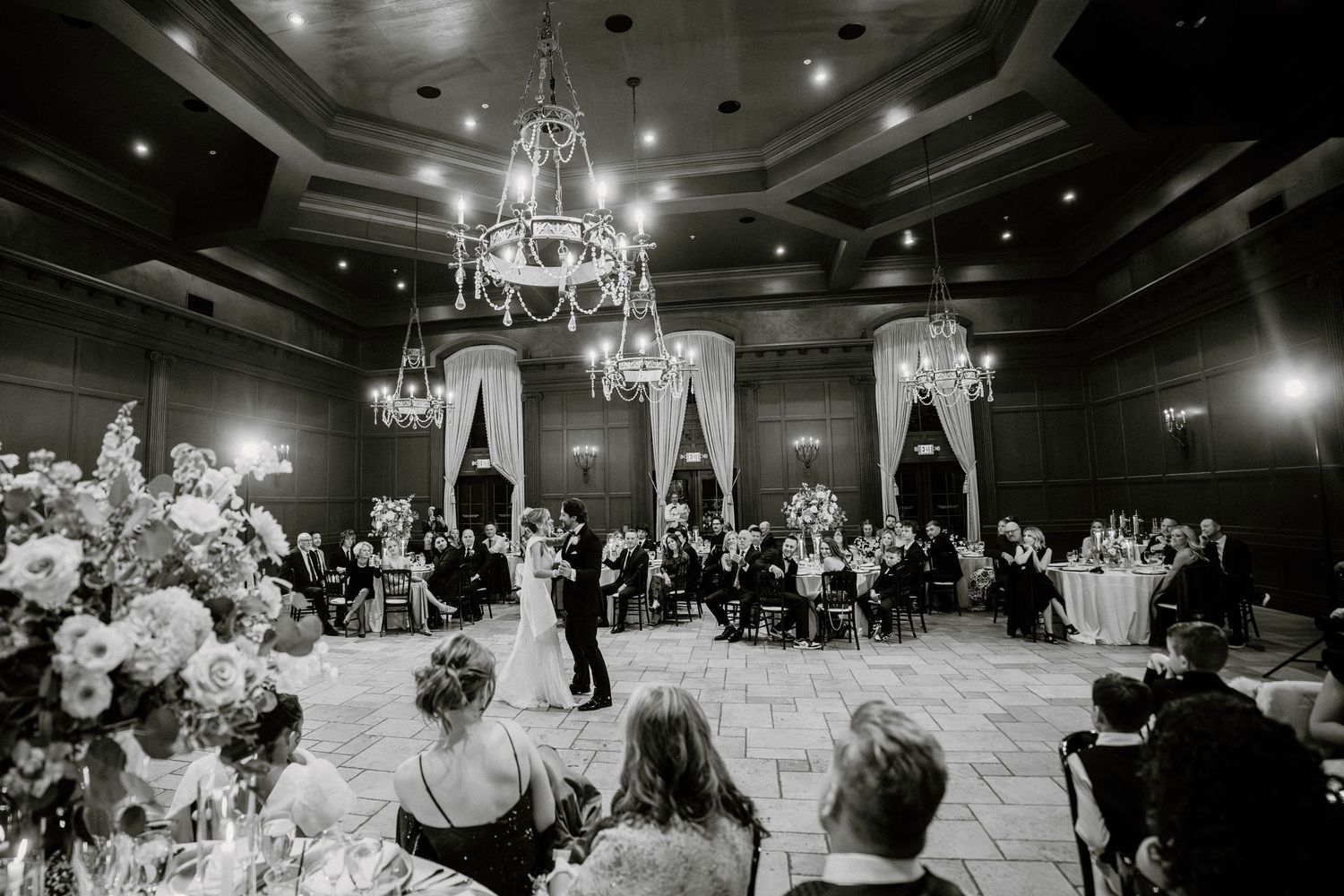 A black and white photograph of an elegant ballroom wedding reception with chandeliers and couples dancing on the floor.