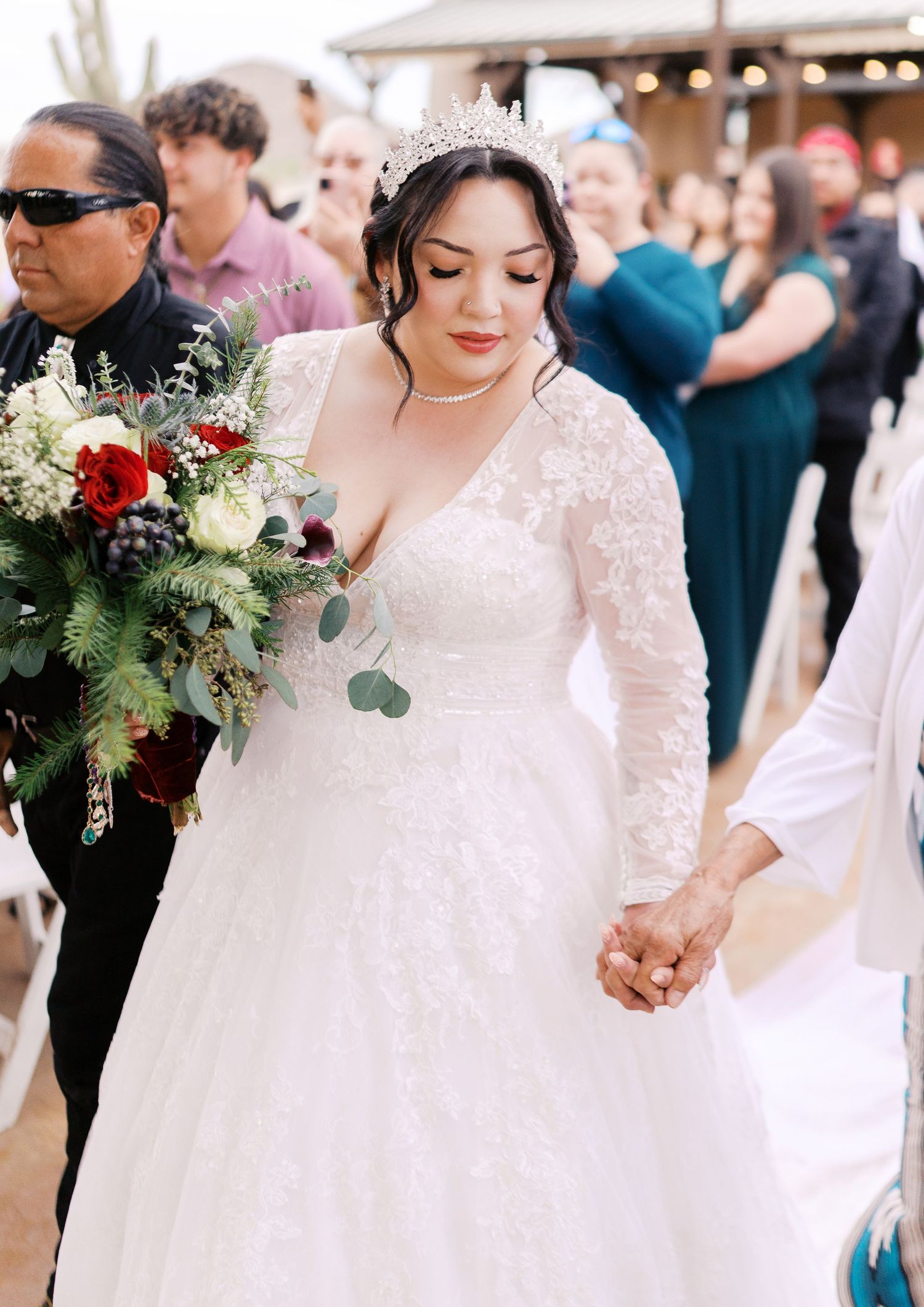 A bride in a long-sleeved lace dress holds a lush bouquet of red and white flowers with eucalyptus at an outdoor wedding.