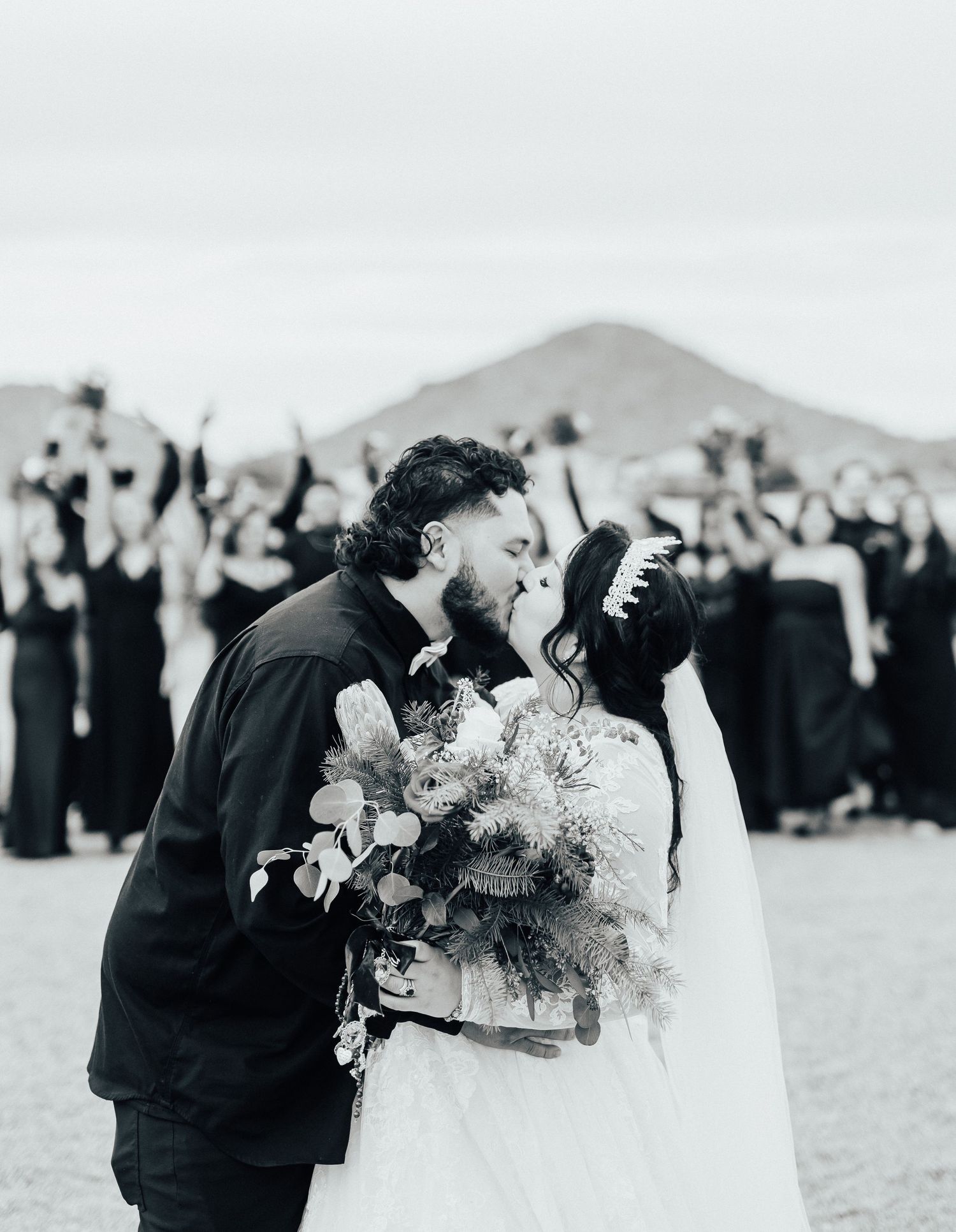 A newly married couple share a romantic kiss outdoors with a mountain in the background and guests watching.