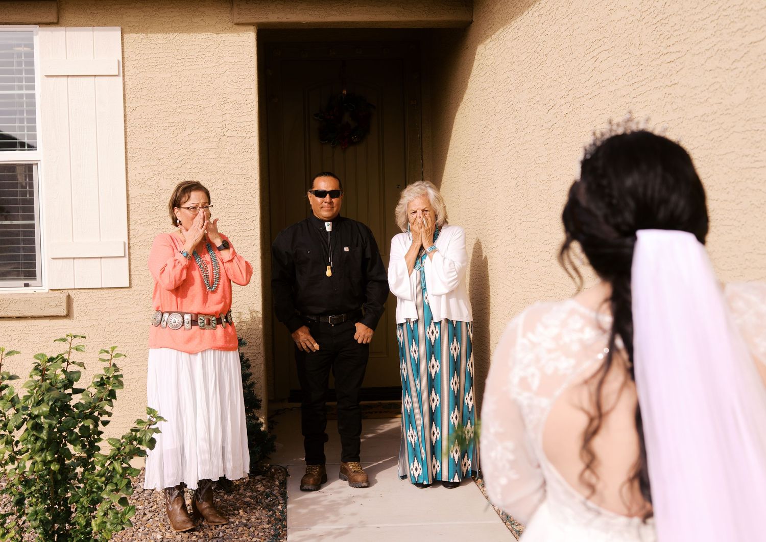 Three people stand in casual and formal attire outside a stucco building entrance with white shutters in daylight.