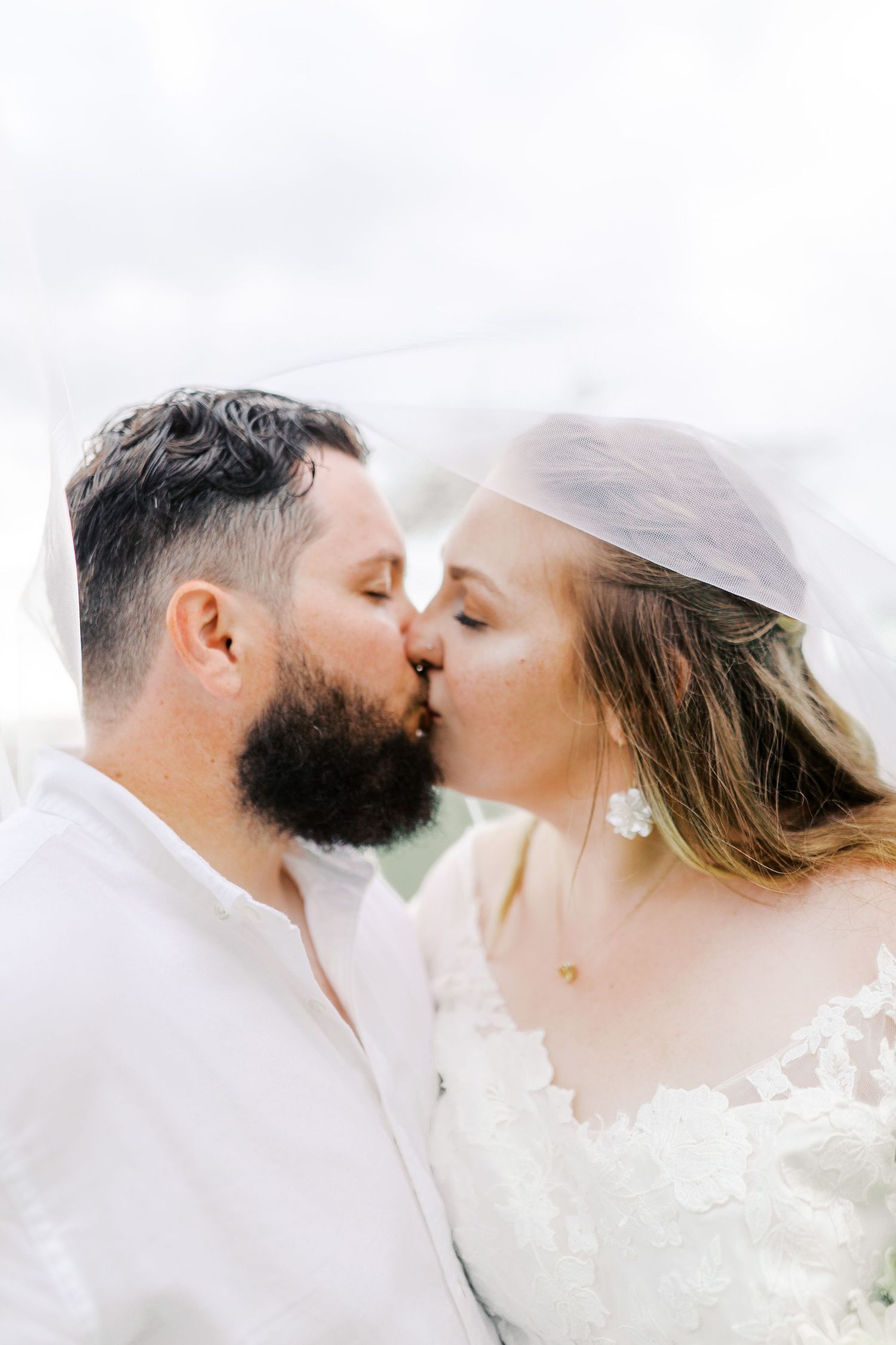 A tender wedding kiss captured in soft natural light with a flowing bridal veil.