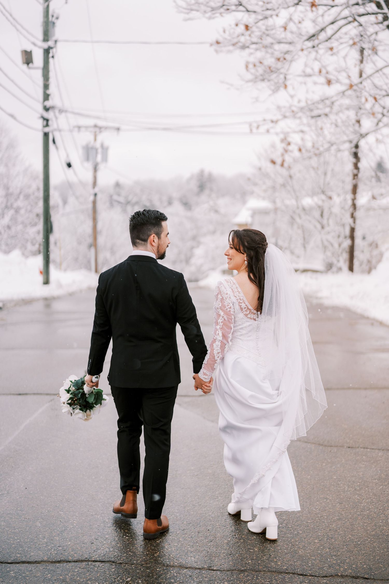 Newlyweds walk hand in hand down a snow-lined street with bride holding a bouquet.
