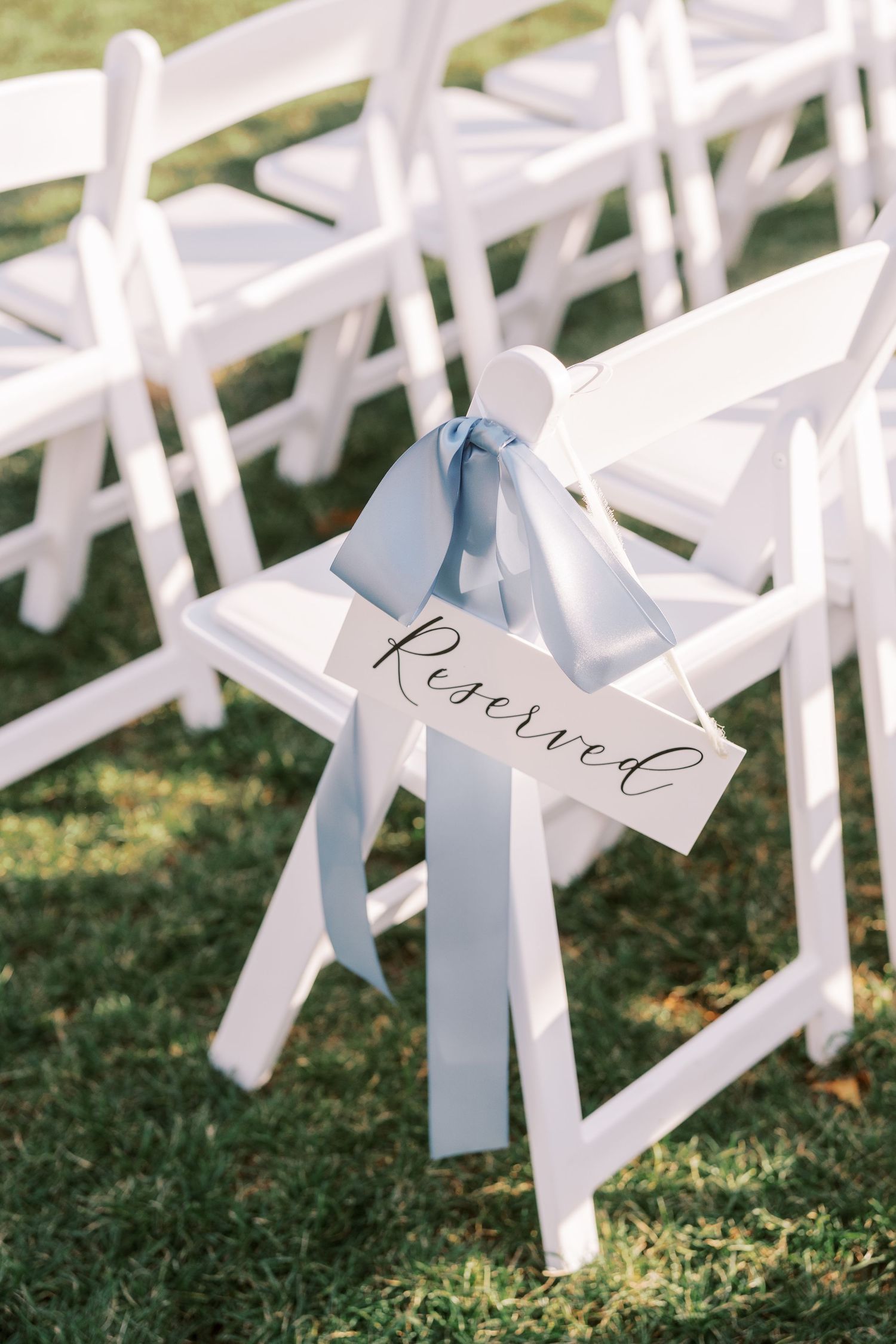 White wedding ceremony chairs with light blue ribbons and name cards arranged on grass for an outdoor celebration.
