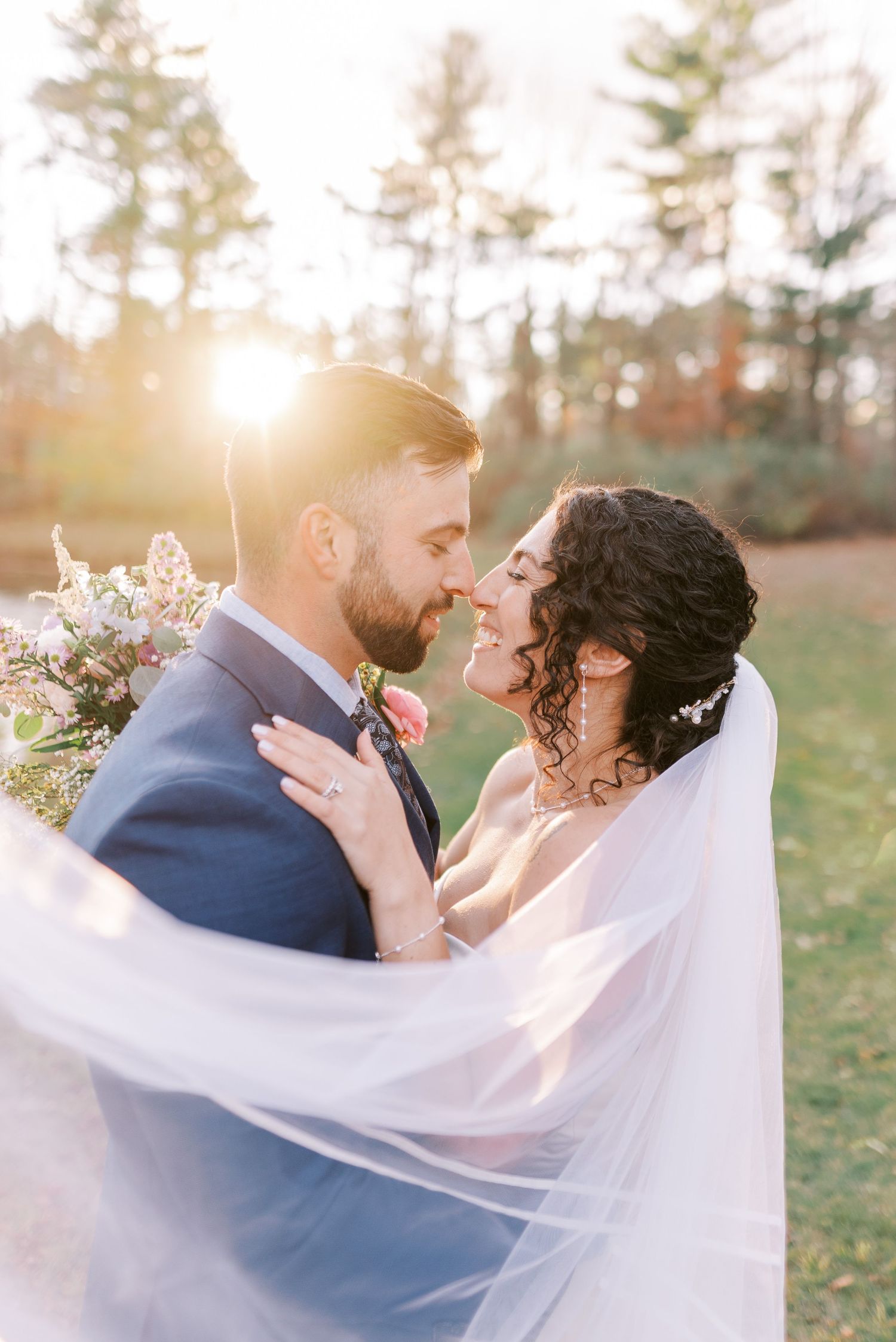 A romantic wedding couple embraces outdoors with a flowing white veil catching the sunlight through trees.