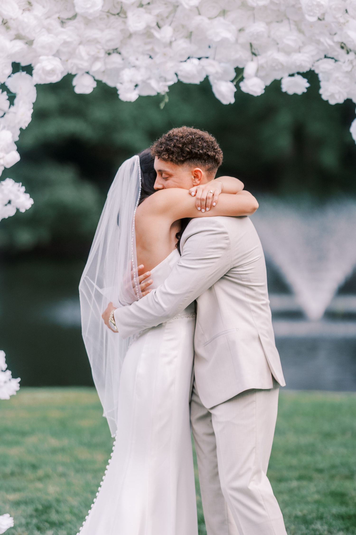 Wedding couple embraces under white floral arch with fountain in background.