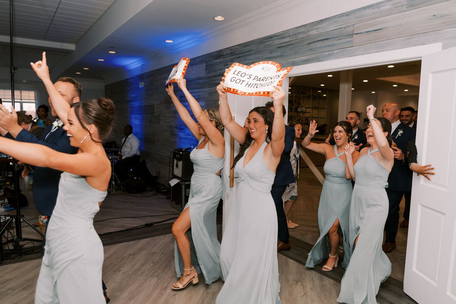 Wedding party members dance in a line with their arms raised at an indoor reception venue with blue lighting.