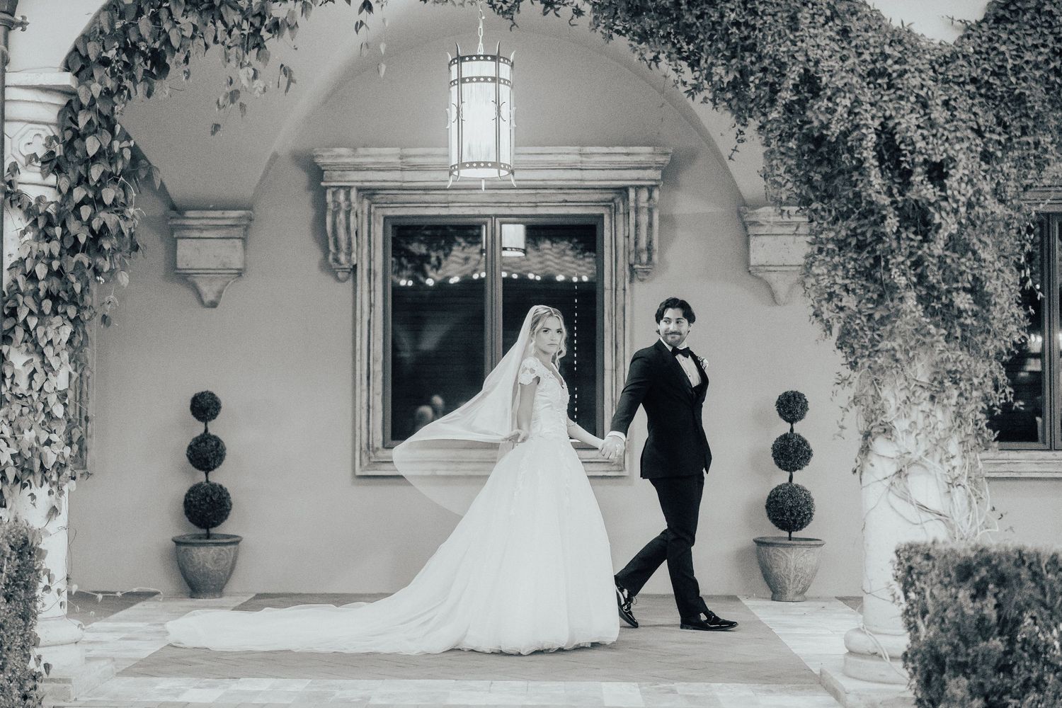 A bride and groom pose together in front of an elegant arched doorway with topiary plants and flowing veil.