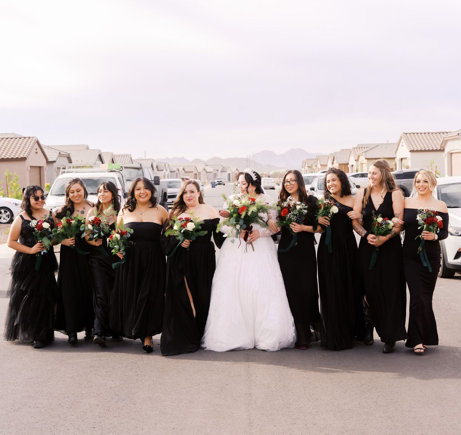A bridal party in black gowns and one white wedding dress pose together outdoors on a suburban street.