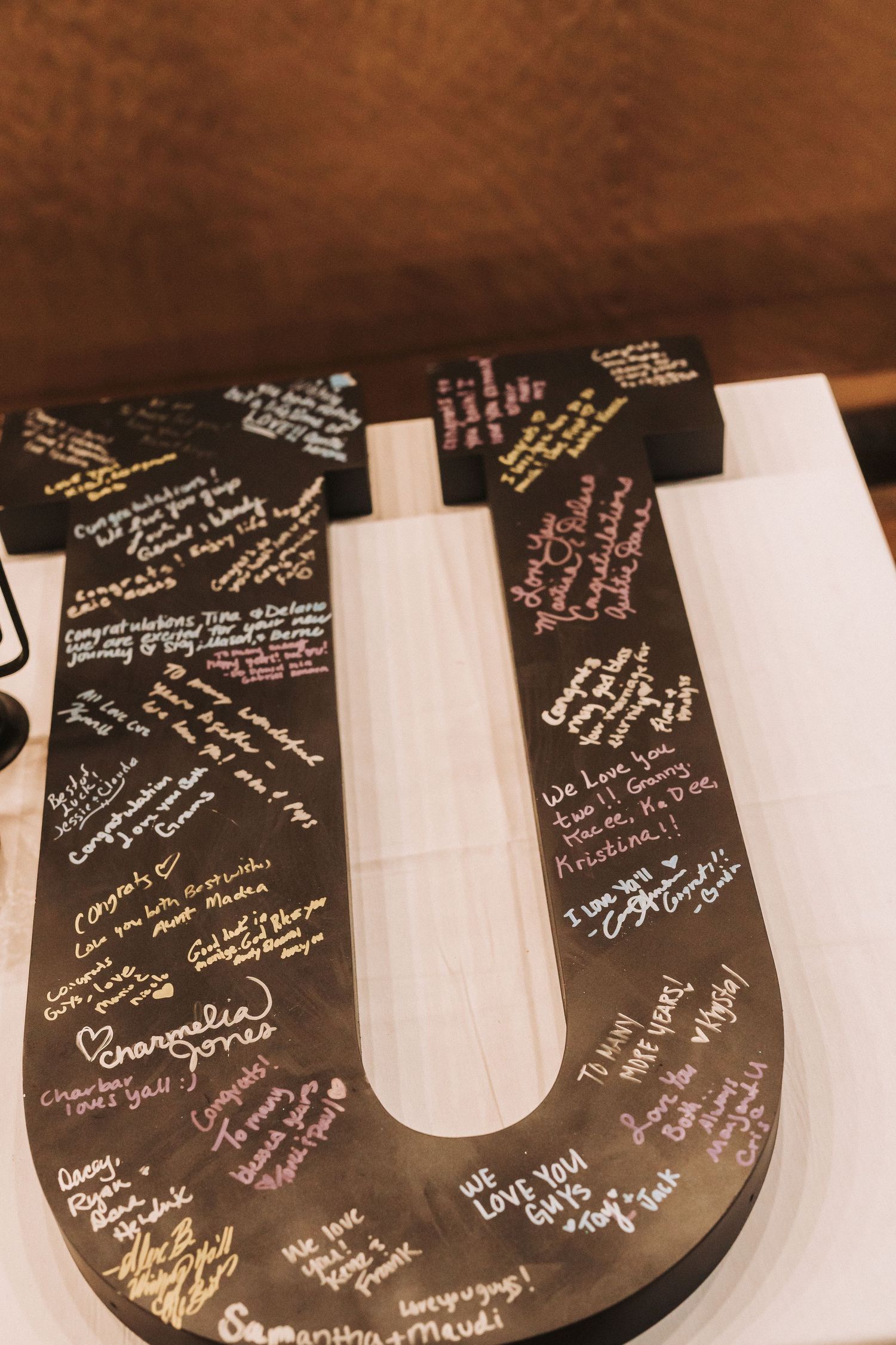 A wooden letter 'U' wedding guest book covered with handwritten signatures and messages in metallic ink against sepia background.