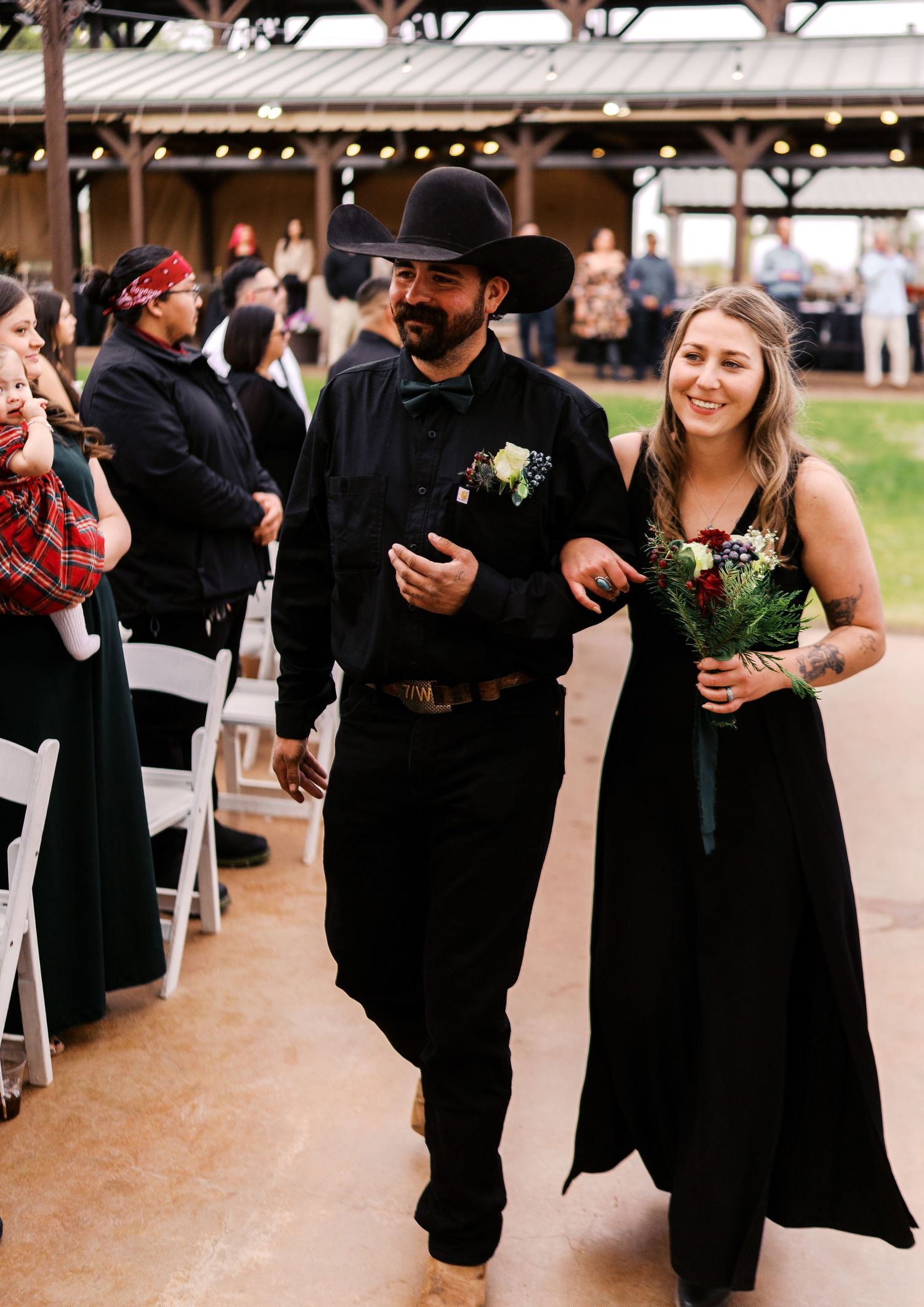 Wedding attendants in black formal attire walk down an aisle at an outdoor covered venue with floral accents.
