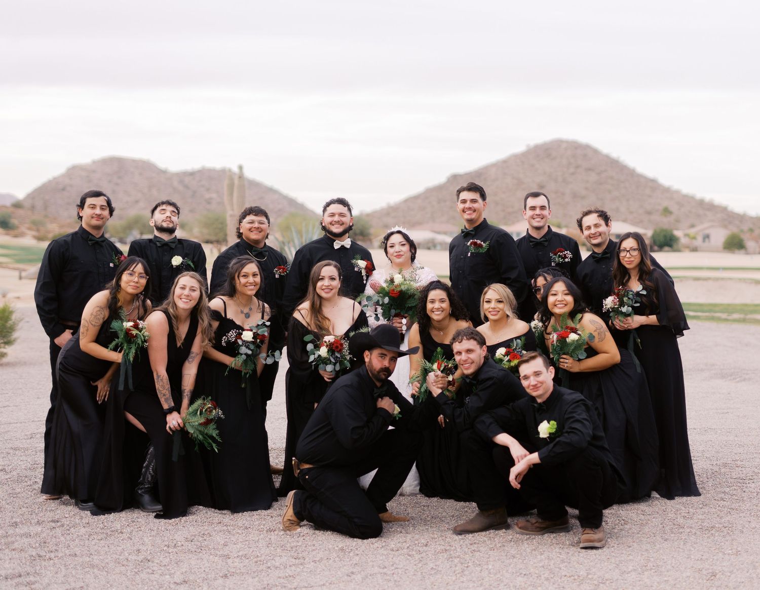 Wedding party in all black formal attire poses together outdoors in desert landscape with mountains in background.