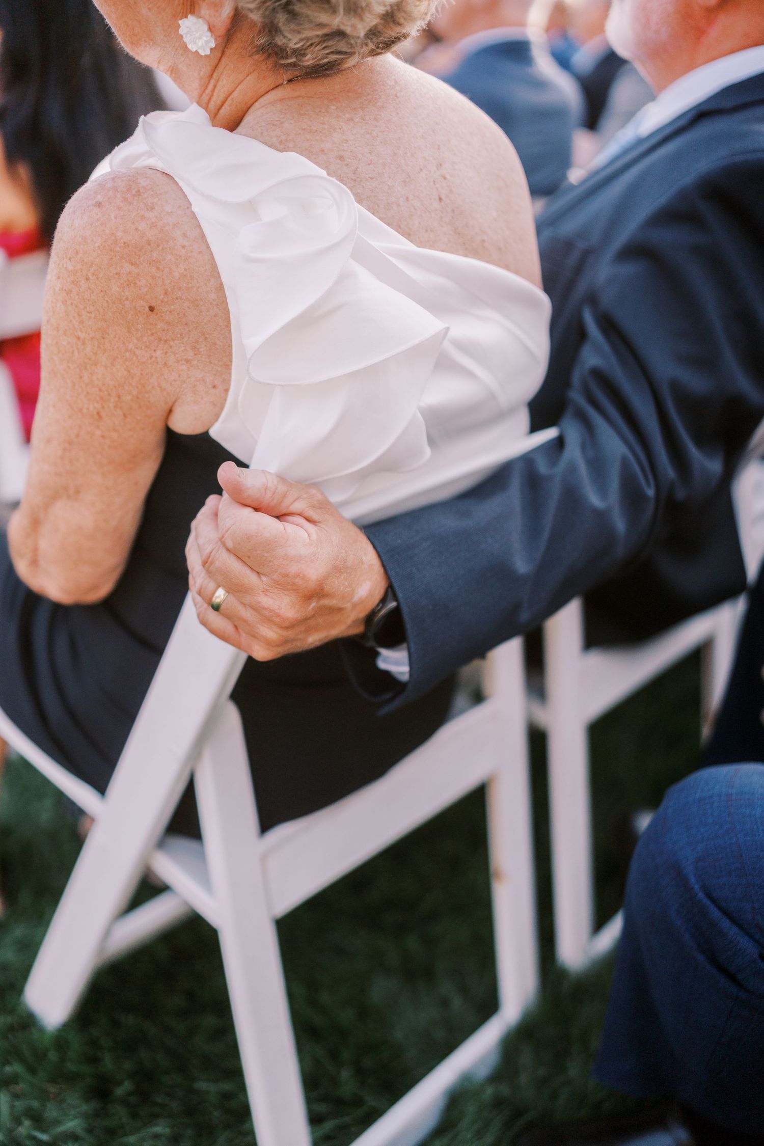 Wedding guests in formal attire seated in white chairs during an outdoor ceremony.
