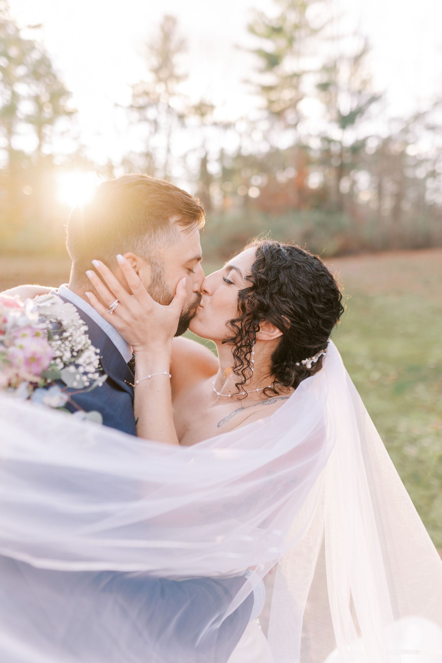 Romantic wedding couple shares intimate kisses in sunlit outdoor setting with flowing white veil in breeze.