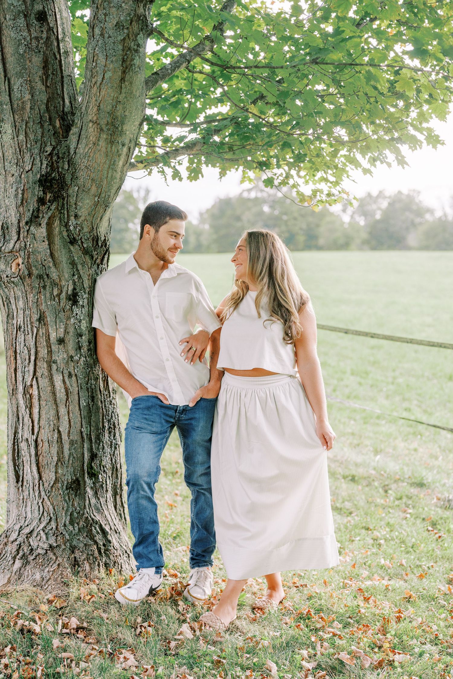 A couple in white clothing poses playfully near large trees during a countryside photo shoot.