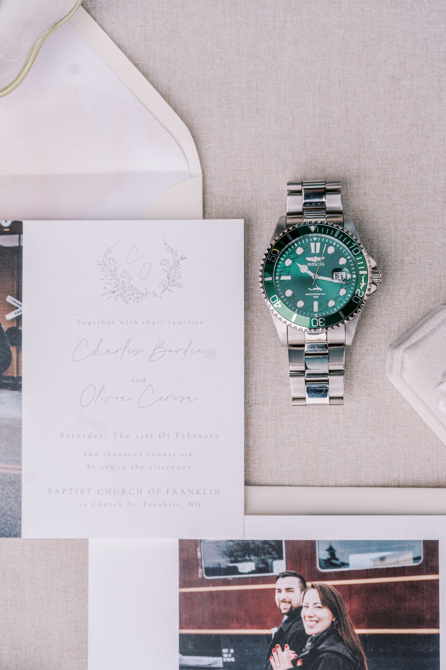 Wedding stationery and a green diving watch arranged on a light grey linen background.