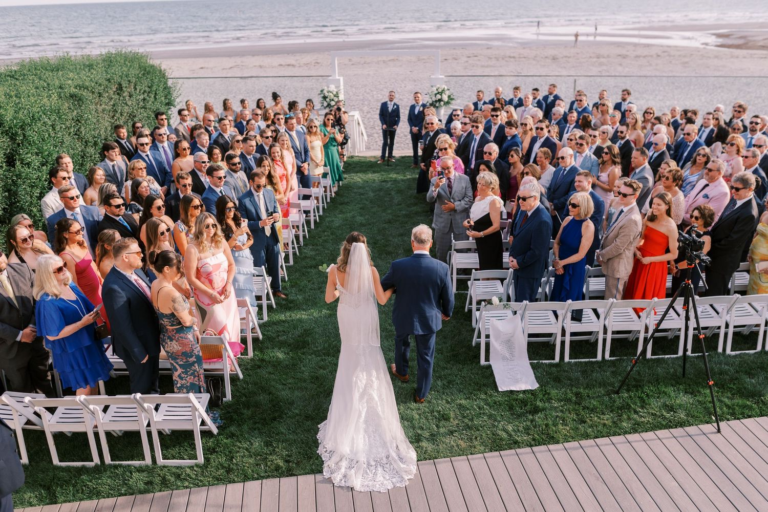A panoramic view of an outdoor wedding ceremony with guests seated in white chairs on a lawn overlooking the ocean.