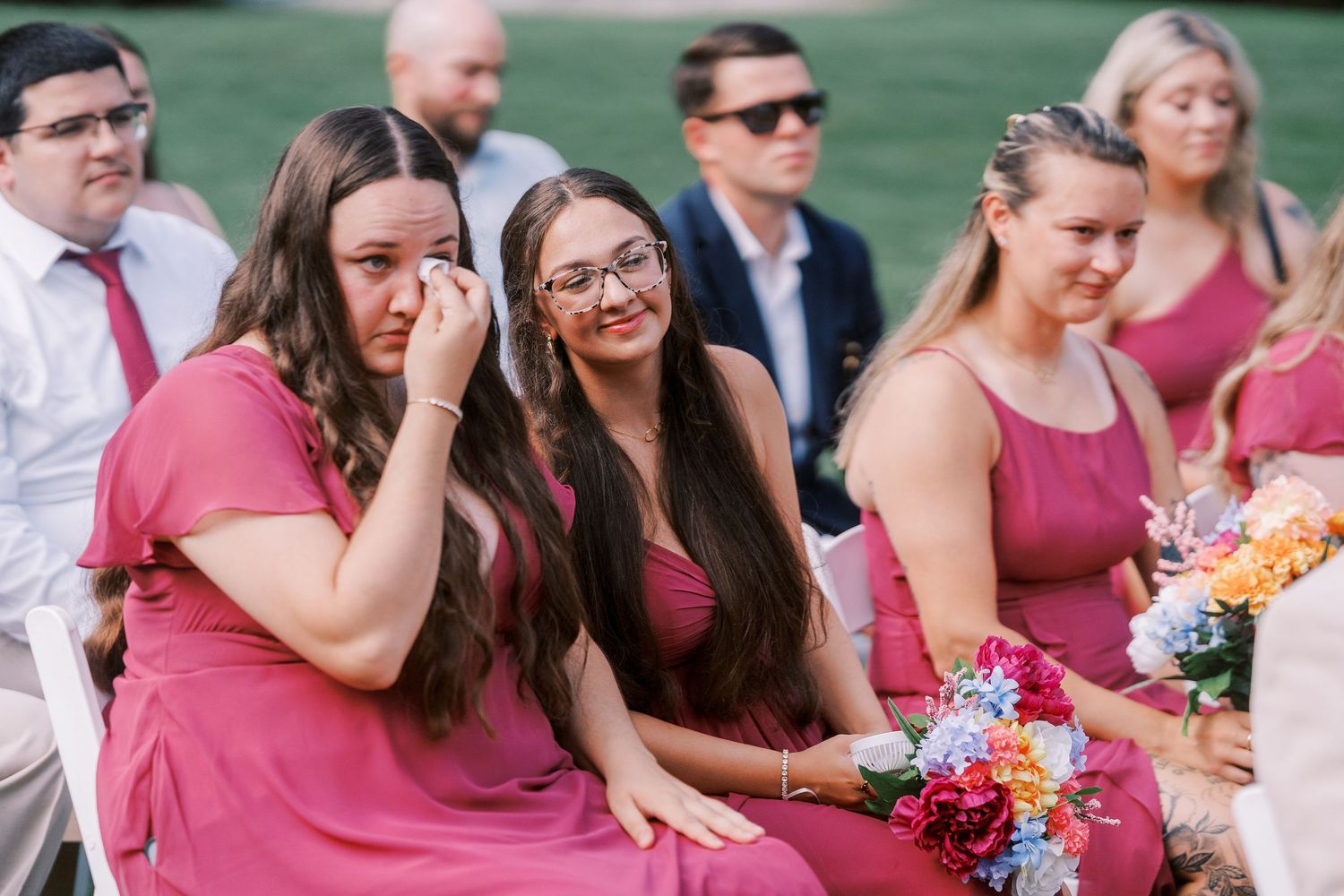 Wedding guests and bridal party in pink dresses seated during outdoor ceremony.