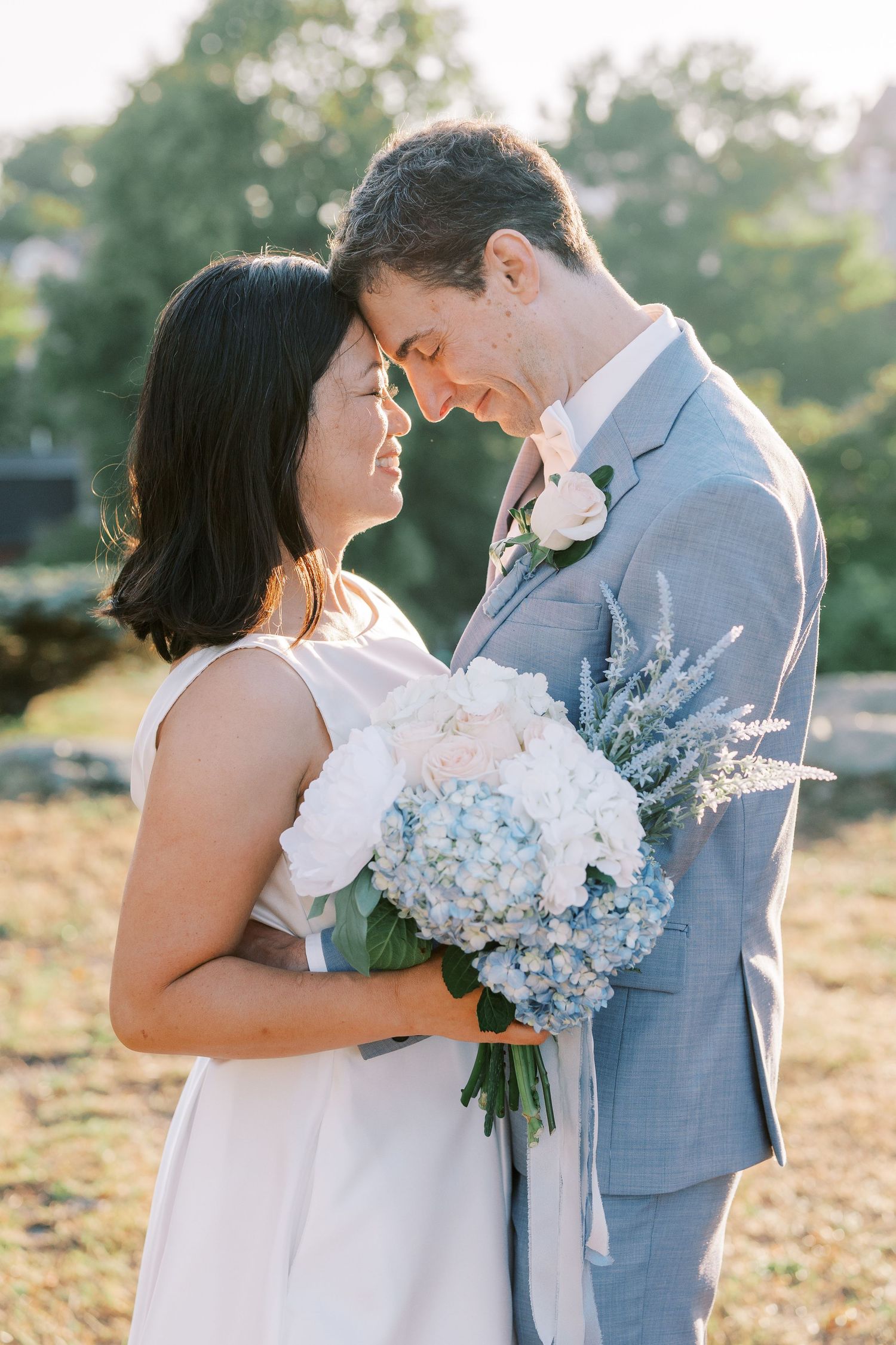 A romantic outdoor wedding scene with a couple embracing while holding light blue hydrangea bouquets.