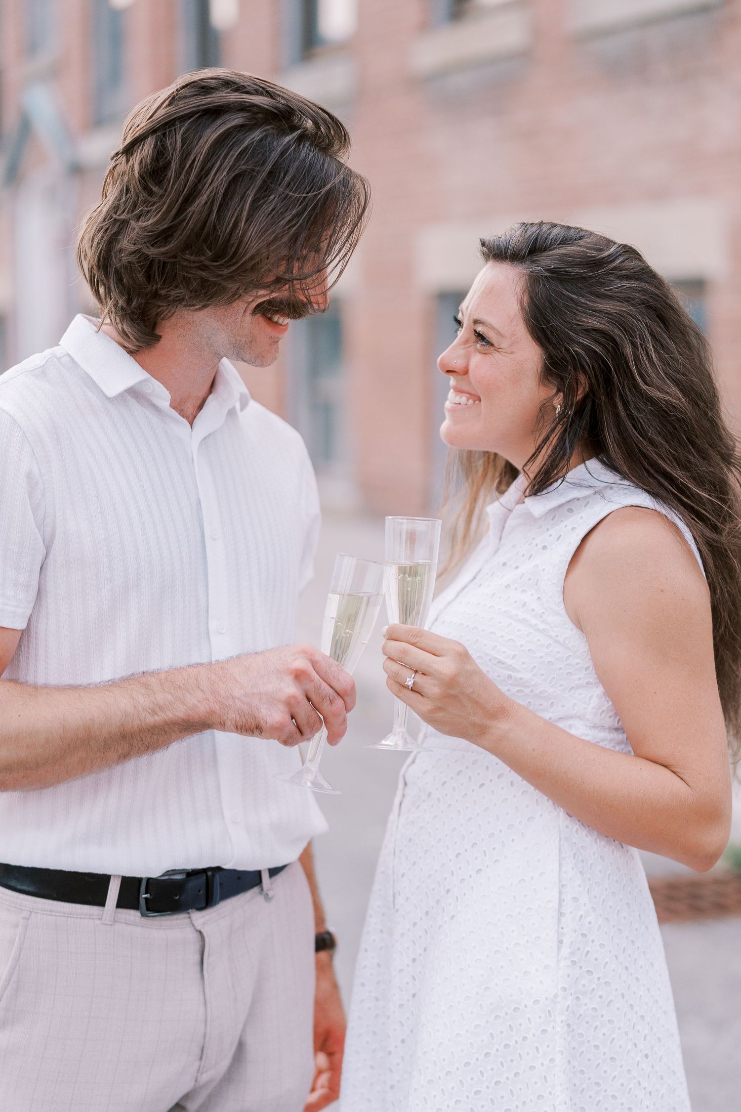 A couple in coordinating white outfits holds champagne glasses while sharing an intimate moment.