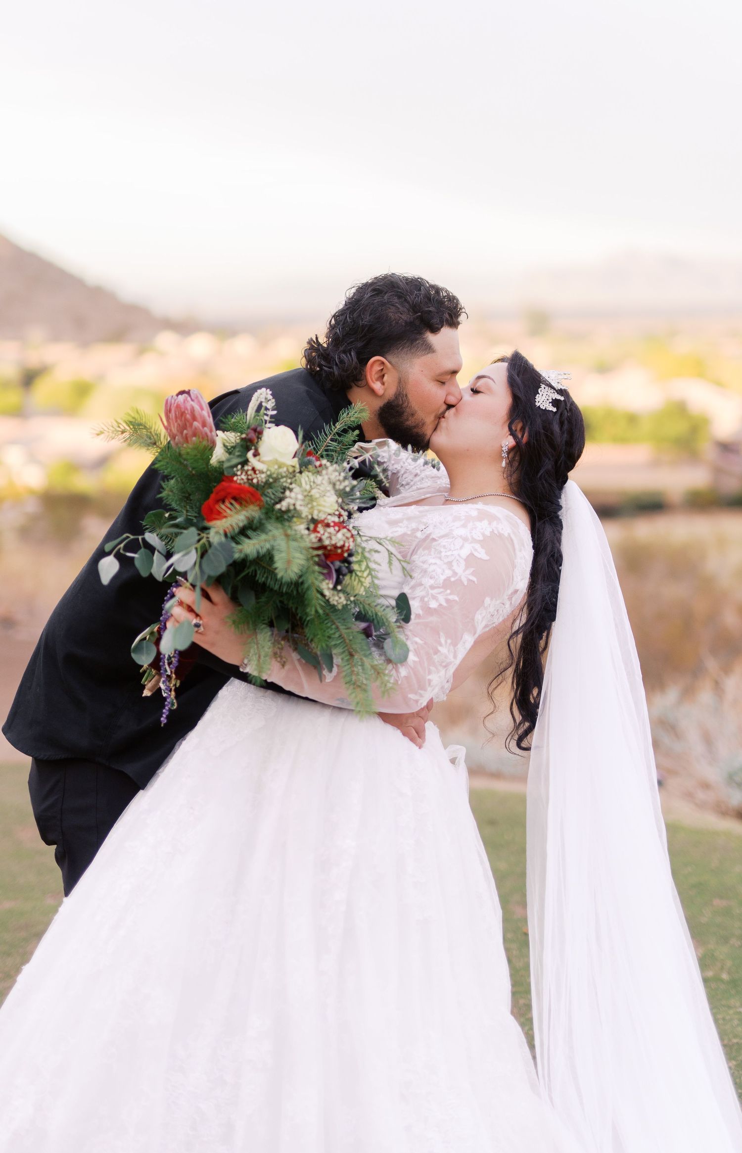 A romantic wedding couple shares a kiss at sunset in a scenic desert landscape while holding a lush floral bouquet.