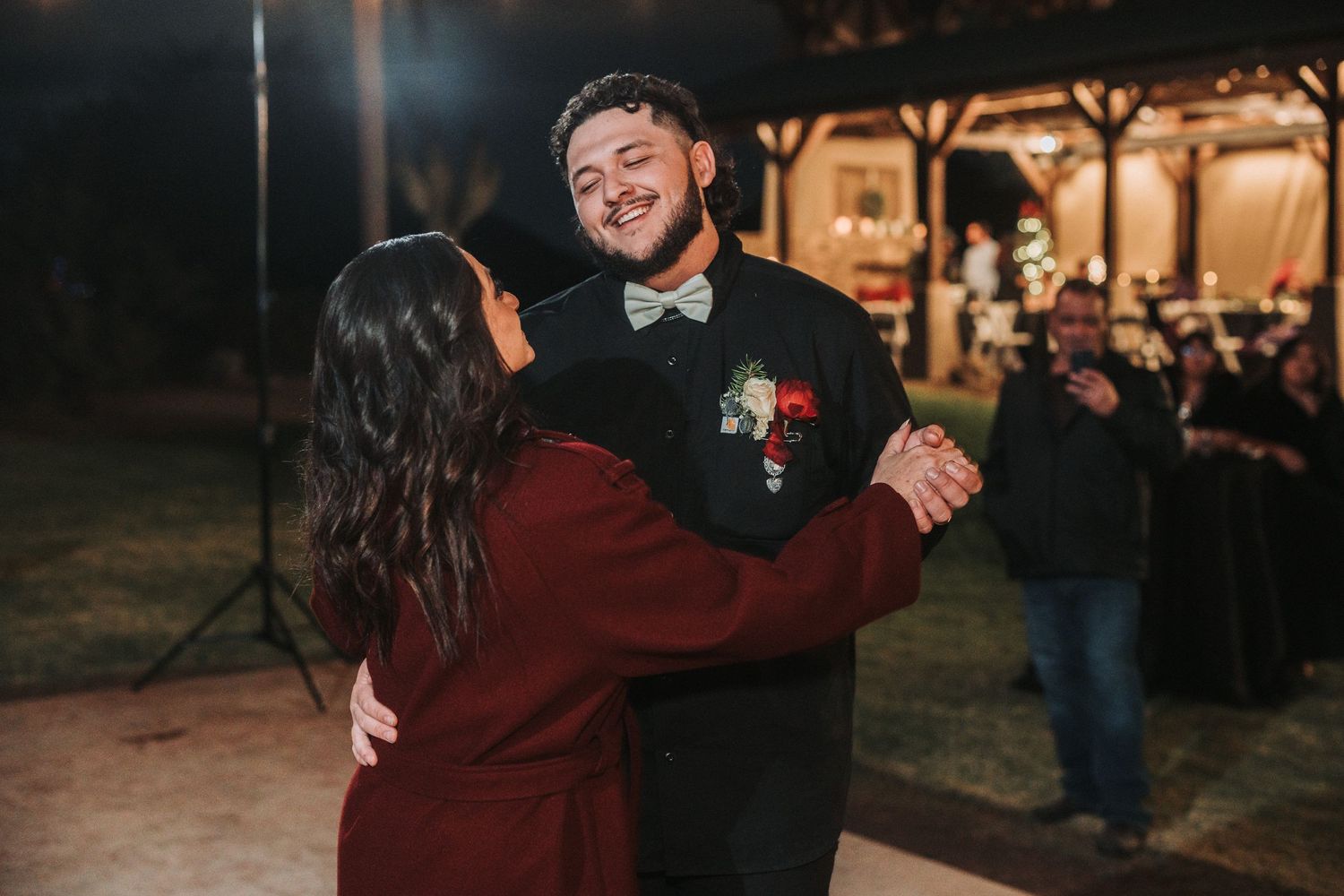A couple dancing together at night under soft lights, with the person in black wearing a boutonniere and their partner in maroon.