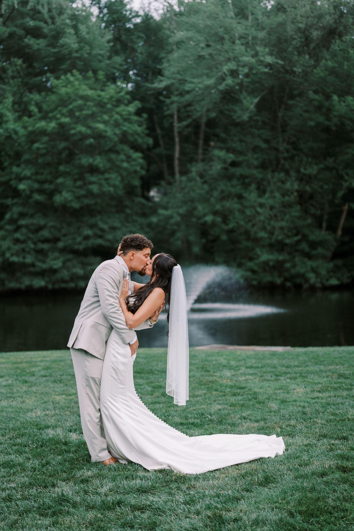 A romantic moment between newlyweds on a lawn with a beautiful pond and fountain in the background.