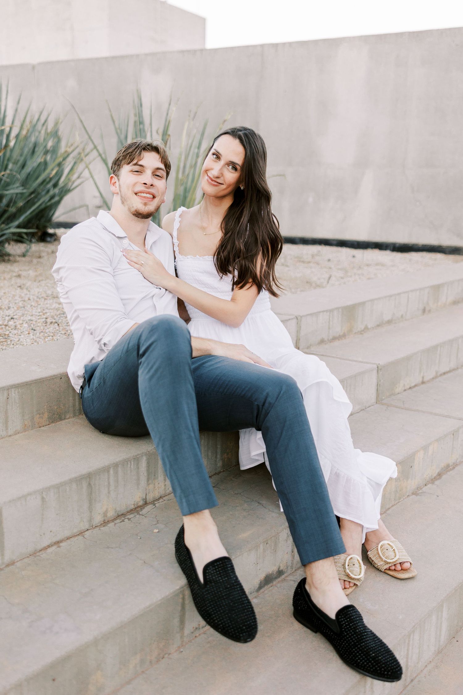 A couple in light colored outfits sitting casually on concrete steps with desert plants in the background.