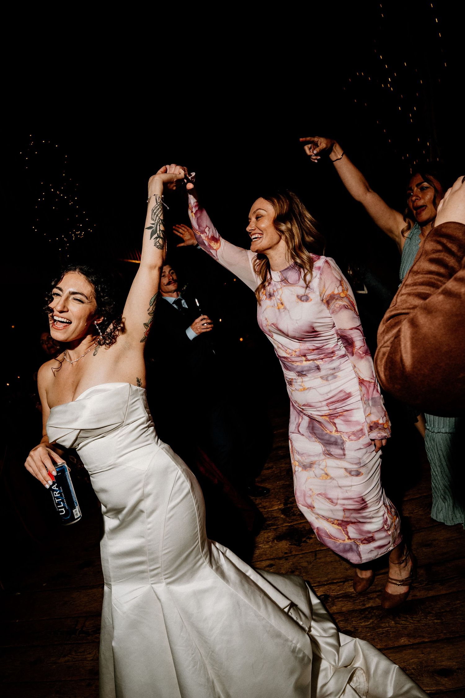 Wedding guests dance energetically on a dark dance floor with joyful expressions and celebratory movements.