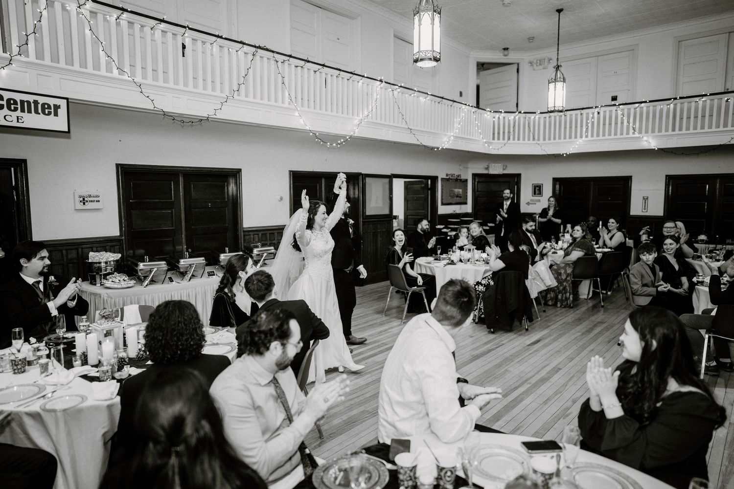 Black and white reception photo showing guests at tables while couple dances on wooden floor.
