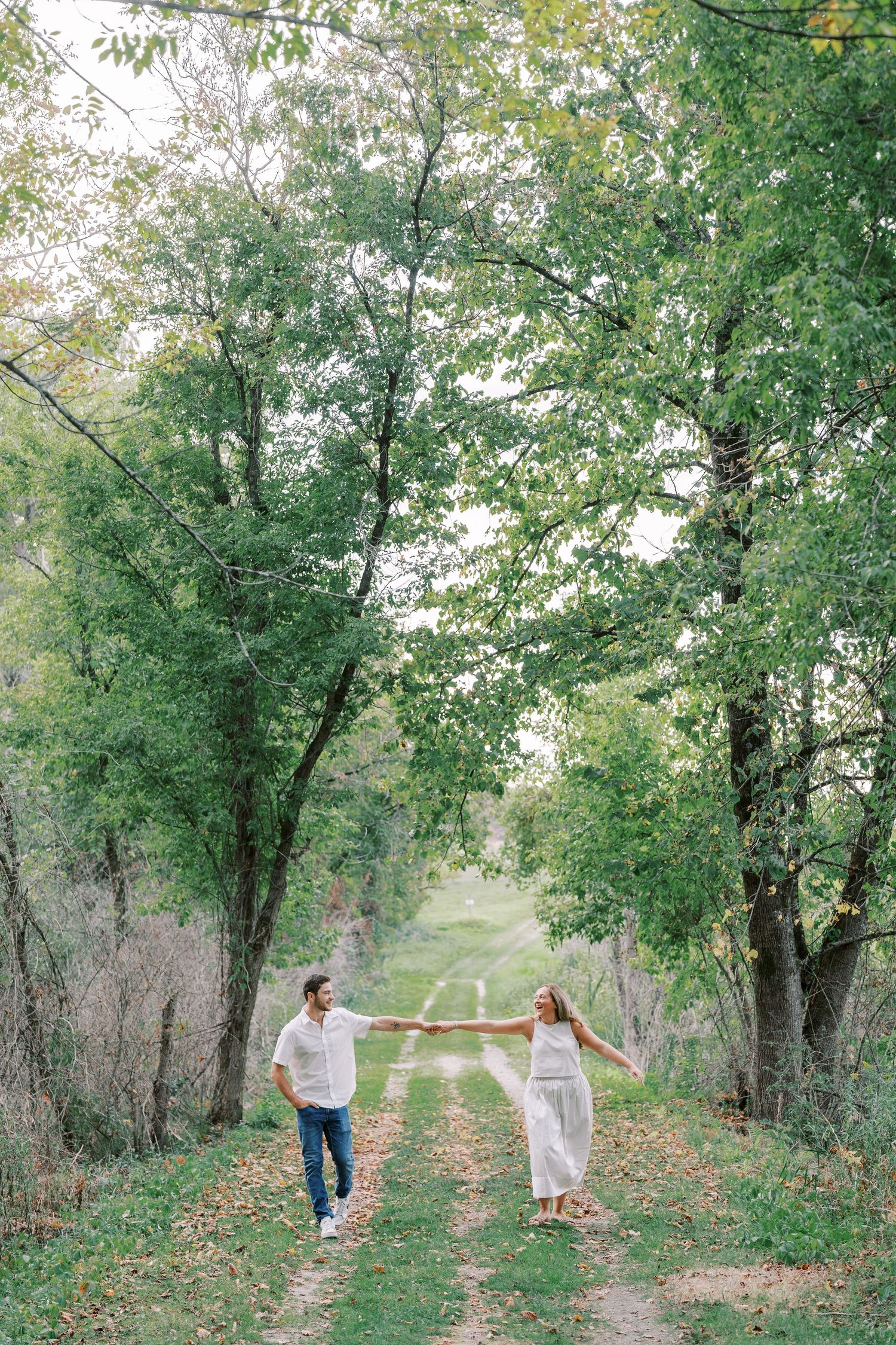 A couple walks hand in hand down a tree-lined path during an outdoor photo session.