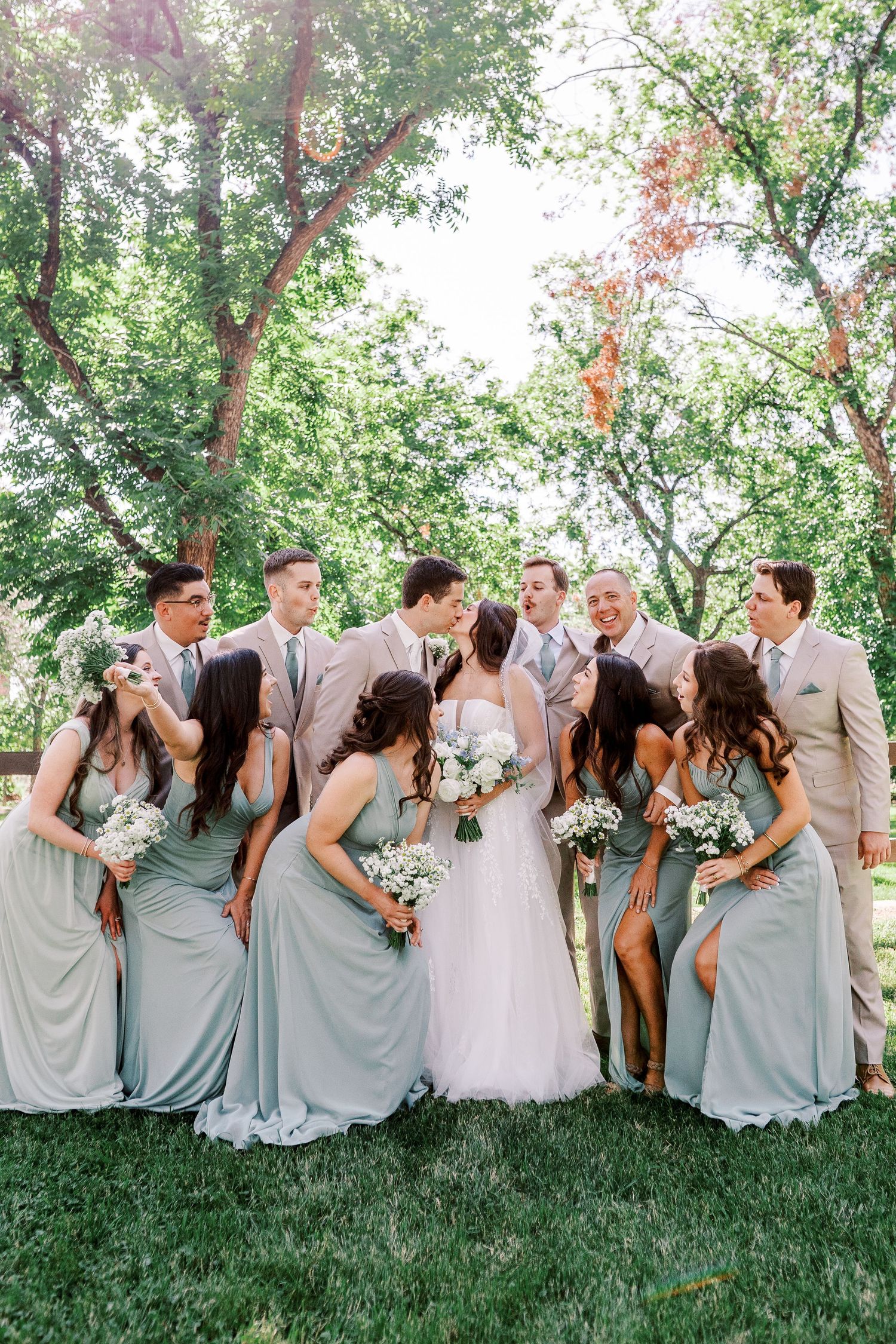 Wedding party in light gray and white attire gather outdoors under green trees for a candid group photo.